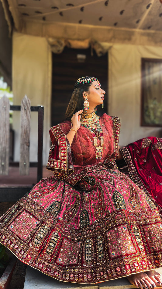 A model wearing Amai Jaipur’s intricately handcrafted red Antriya bridal lehenga, embellished with heavy zari, gota patti, and traditional Rajasthani embroidery, styled with ornate gold and multicolor gemstone jewelry, captured in a royal outdoor setting.