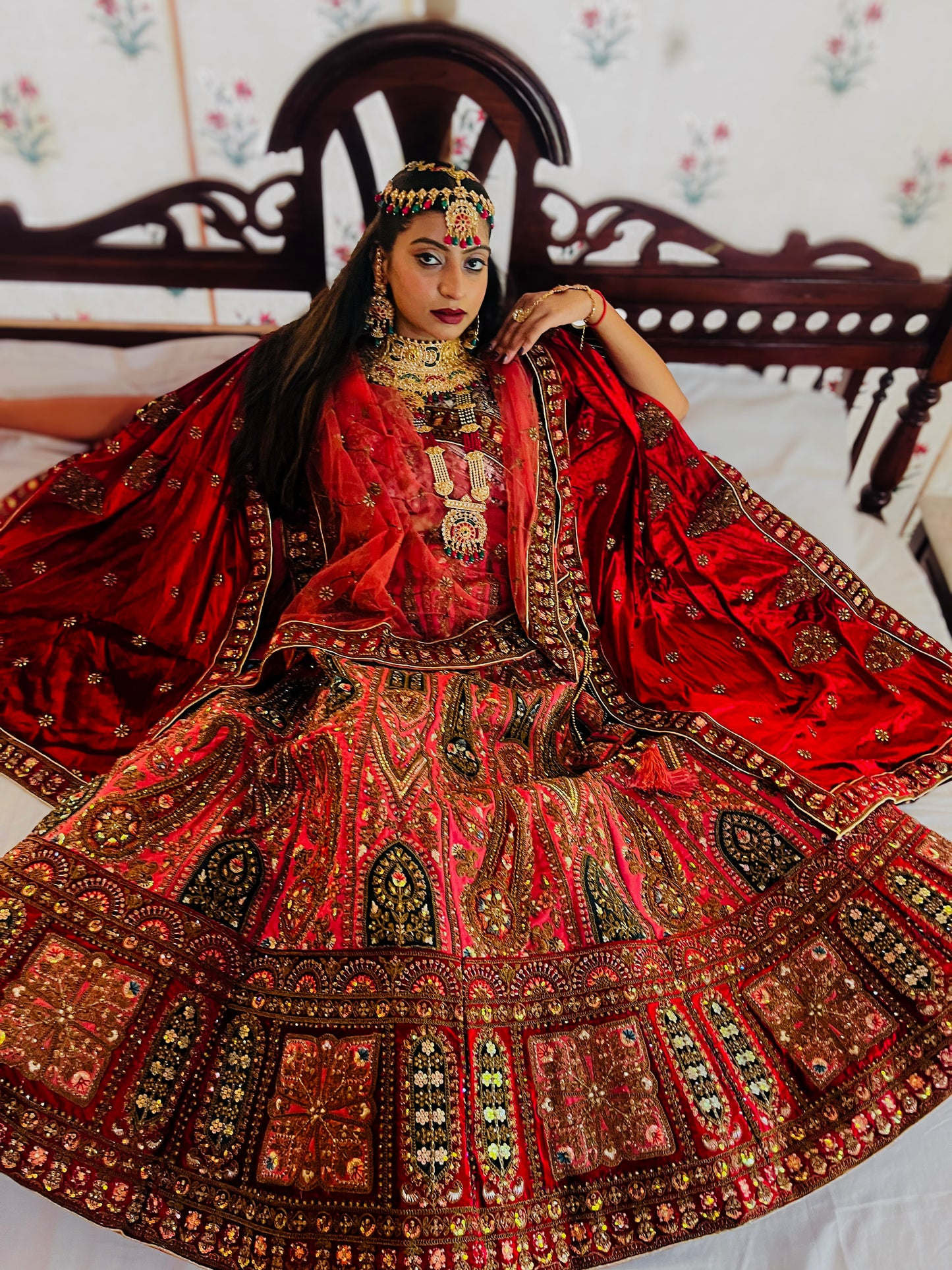 A woman dressed in an ornate red traditional lehenga featuring heavy gold embroidery, mirror work, and intricate motifs, paired with elaborate kundan jewelry and a matching dupatta, posing gracefully on a carved wooden bed; showcasing luxurious Indian bridal fashion craftsmanship.