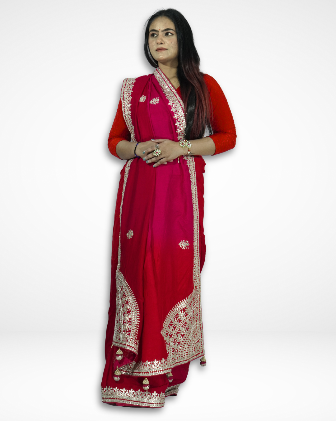Woman modeling a vibrant red saree with intricate gold embroidery along the border and pallu, paired with a red blouse and traditional jewelry, standing against a plain background. ✨