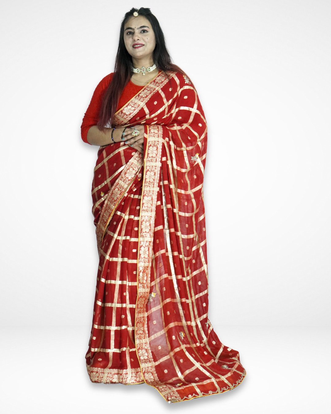 Model wearing a rich red saree adorned with a gold checkered pattern, delicate buti motifs, and an ornate zari border, styled with a matching blouse and statement jewelry for a regal festive look by Amai Jaipur. ✨