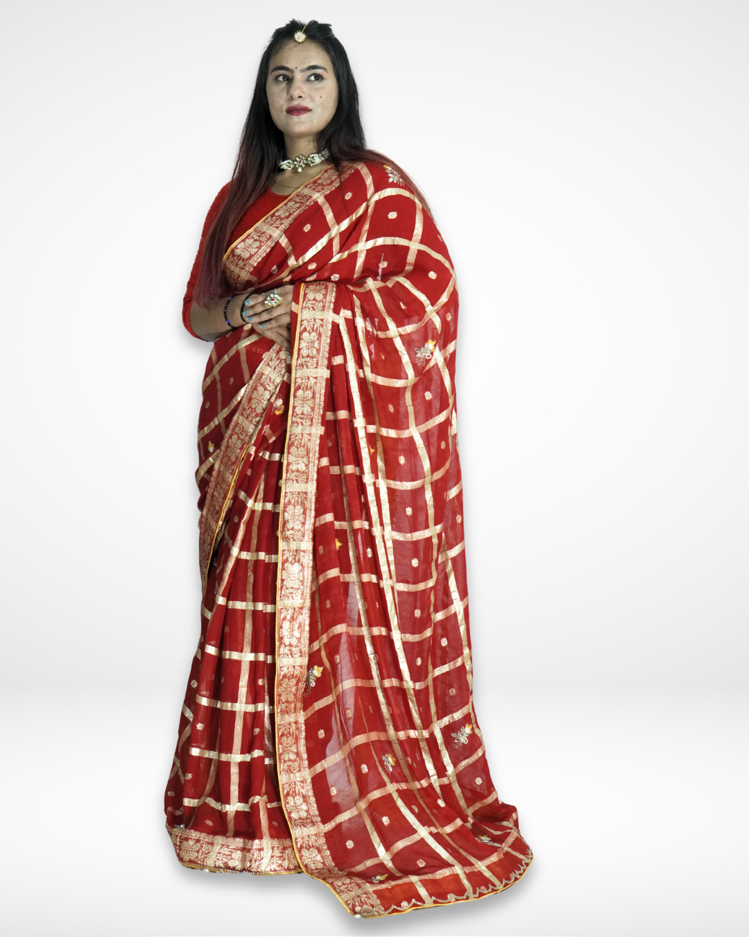 Model draped in a luxurious red saree featuring a gold checkered pattern with delicate buti motifs and an intricate zari border, styled with a matching blouse and statement jewelry for a regal festive look by Amai Jaipur. ✨