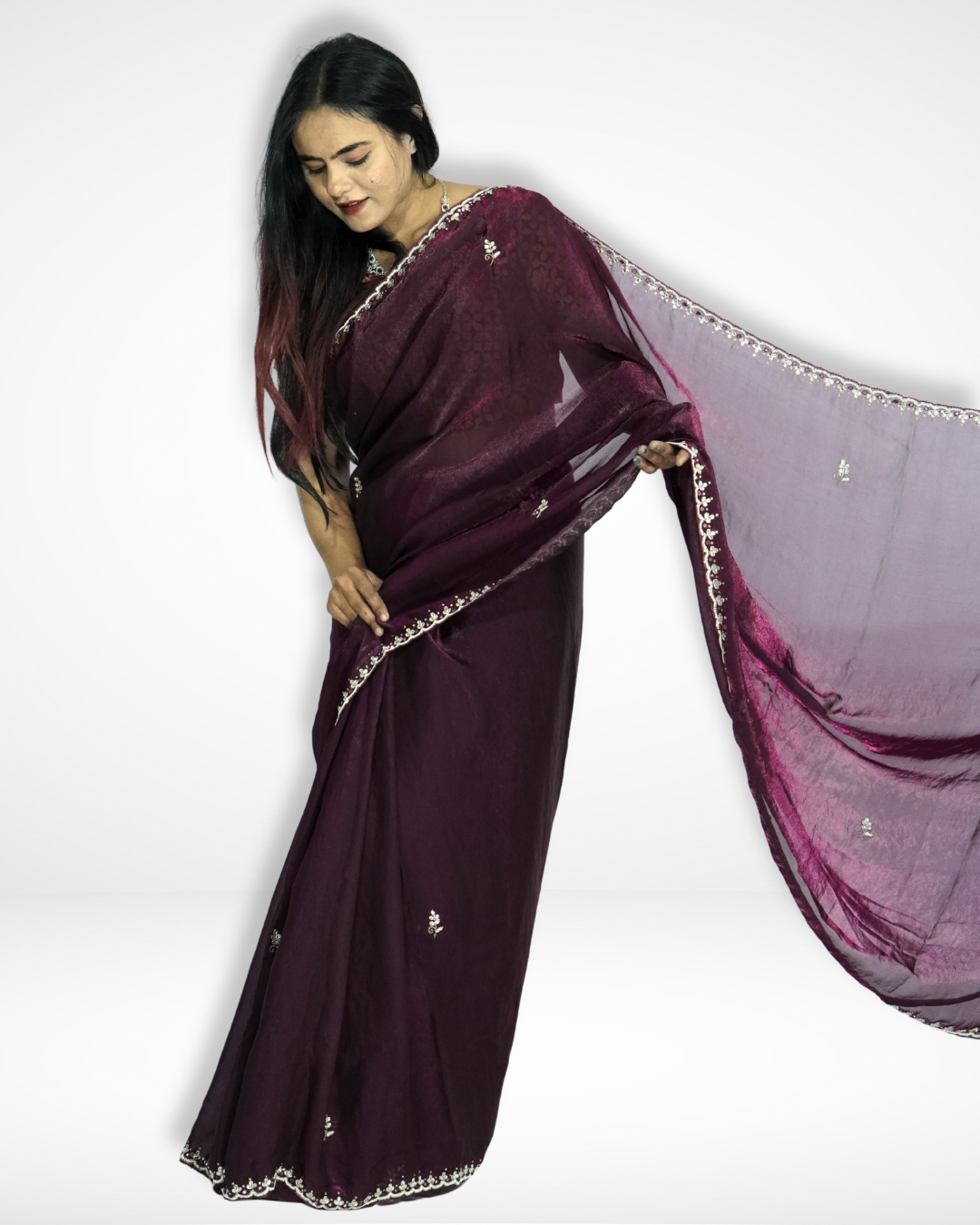 Woman posing in a deep wine-purple saree with a soft shimmer, delicately embroidered border, and scattered motifs, gracefully extending the flowing pallu to showcase the elegant drape. ✨