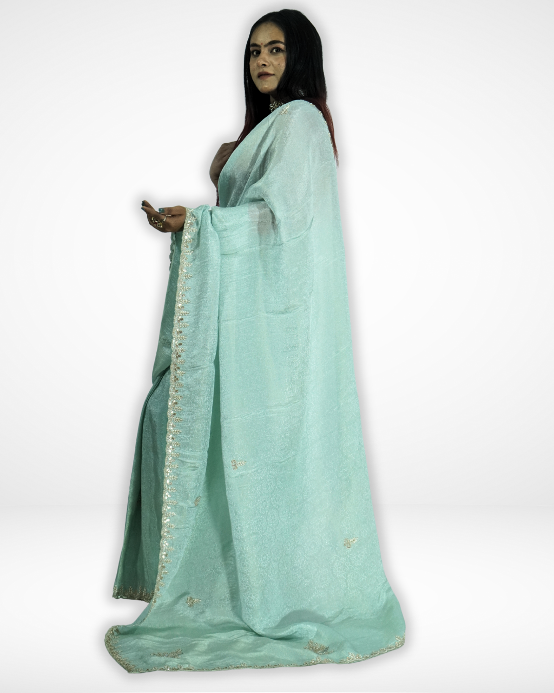 Side view of a woman draped in a mint green saree with delicate gold embroidered borders and small motifs, paired with a red blouse and traditional jewelry, showcasing the flowing pallu against a plain background. ✨