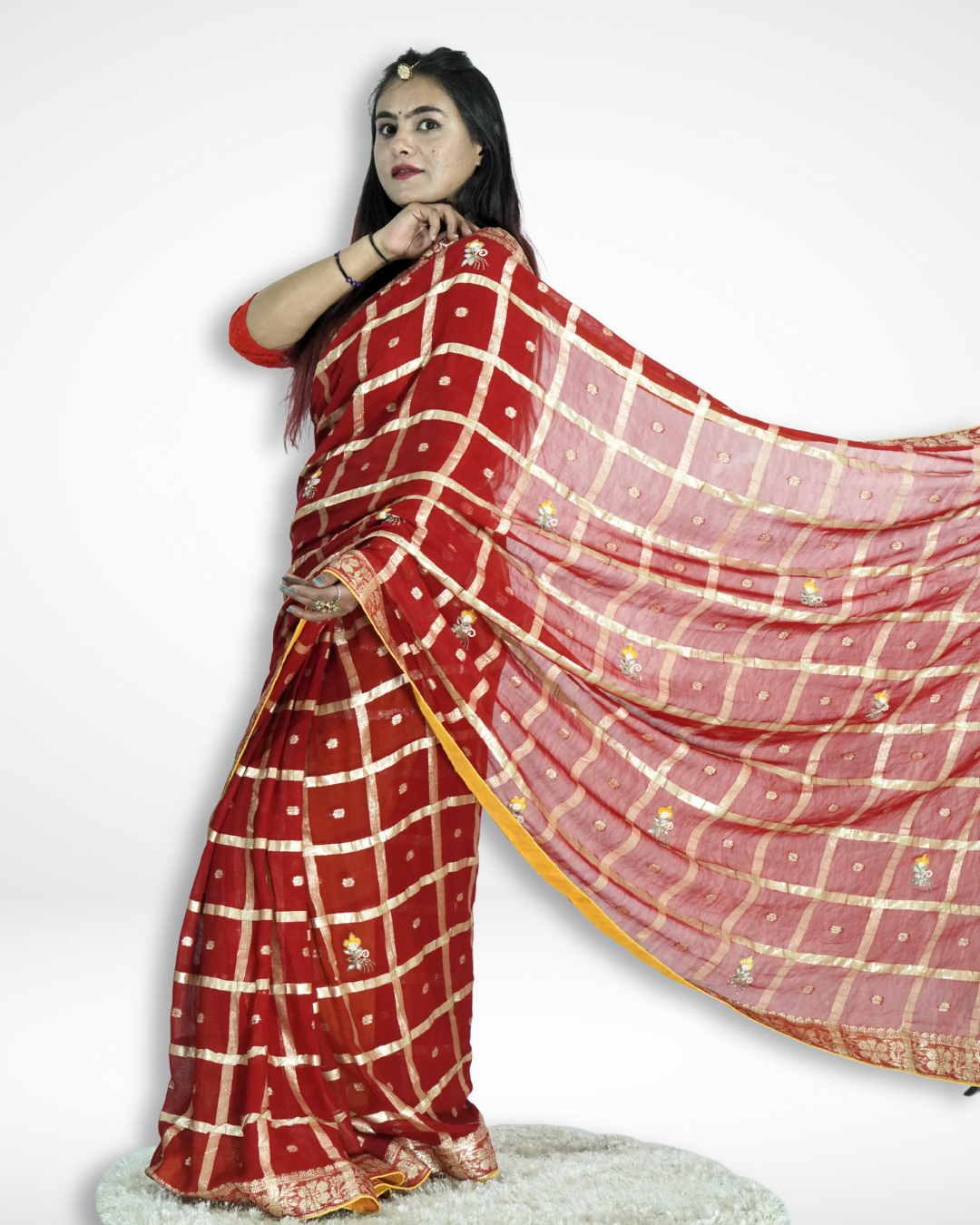 Model showcasing a striking red saree with a gold checkered pattern and delicate buti motifs, featuring a flowing pallu and ornate zari border, styled for a graceful festive look by Amai Jaipur. ✨