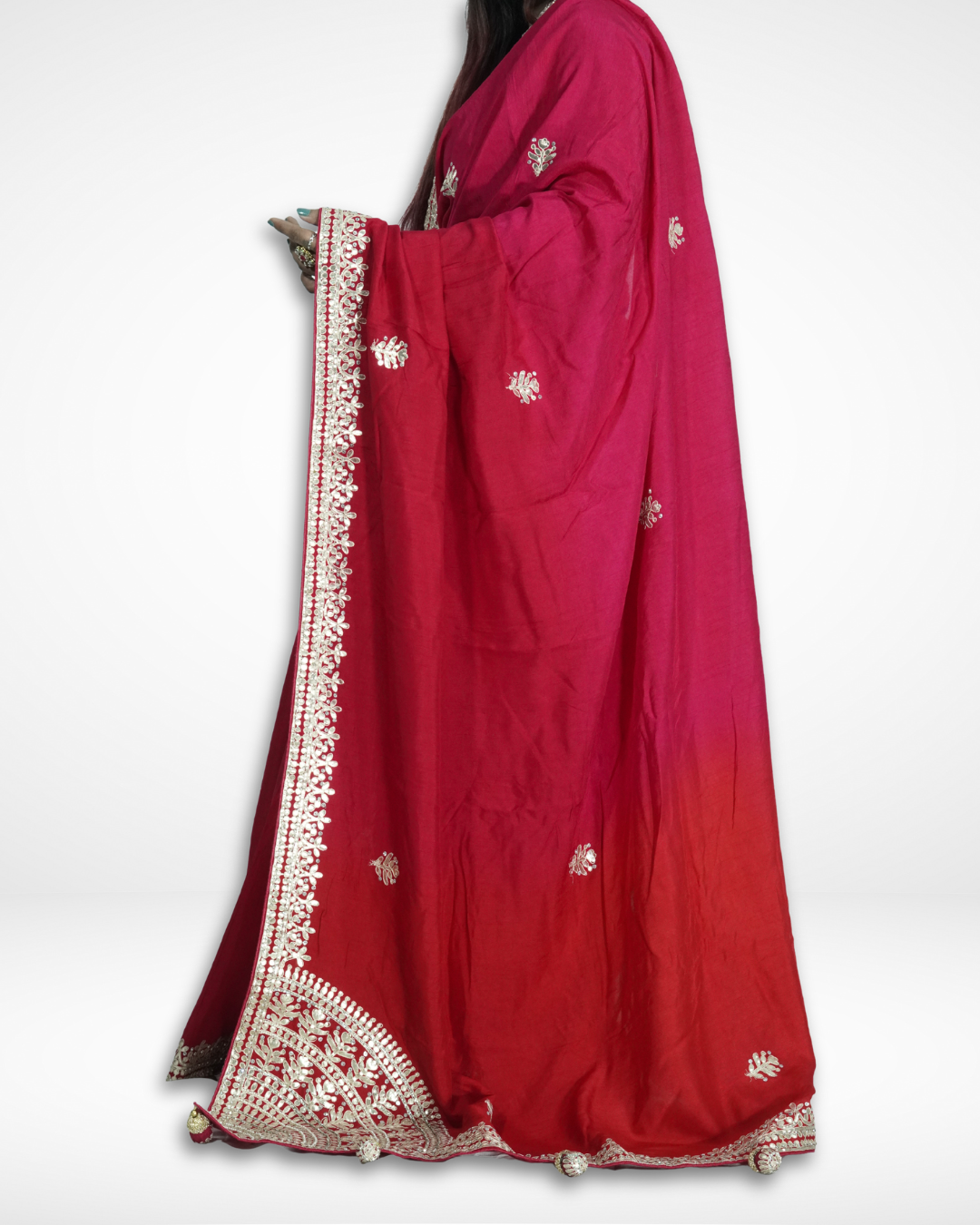 Side view of a woman draped in a red saree featuring delicate gold embroidered borders, small floral motifs across the fabric, and an ornate corner pallu design with tassel accents, styled against a plain background. ✨