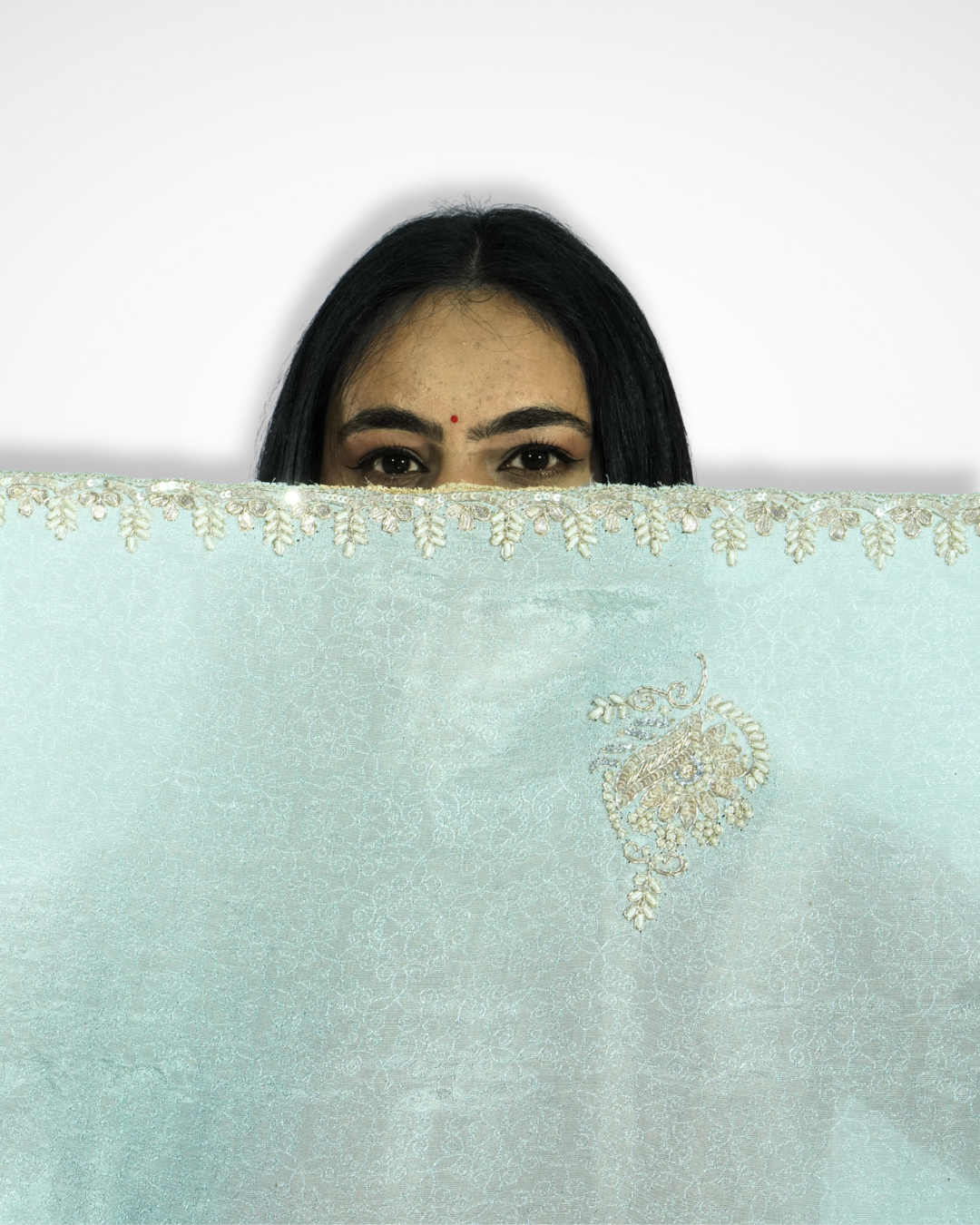 Close-up of a mint green saree with delicate gold embroidered border and paisley motif, with a woman partially hiding behind the fabric so that only her eyes are visible above the saree, creating an elegant and playful pose. ✨