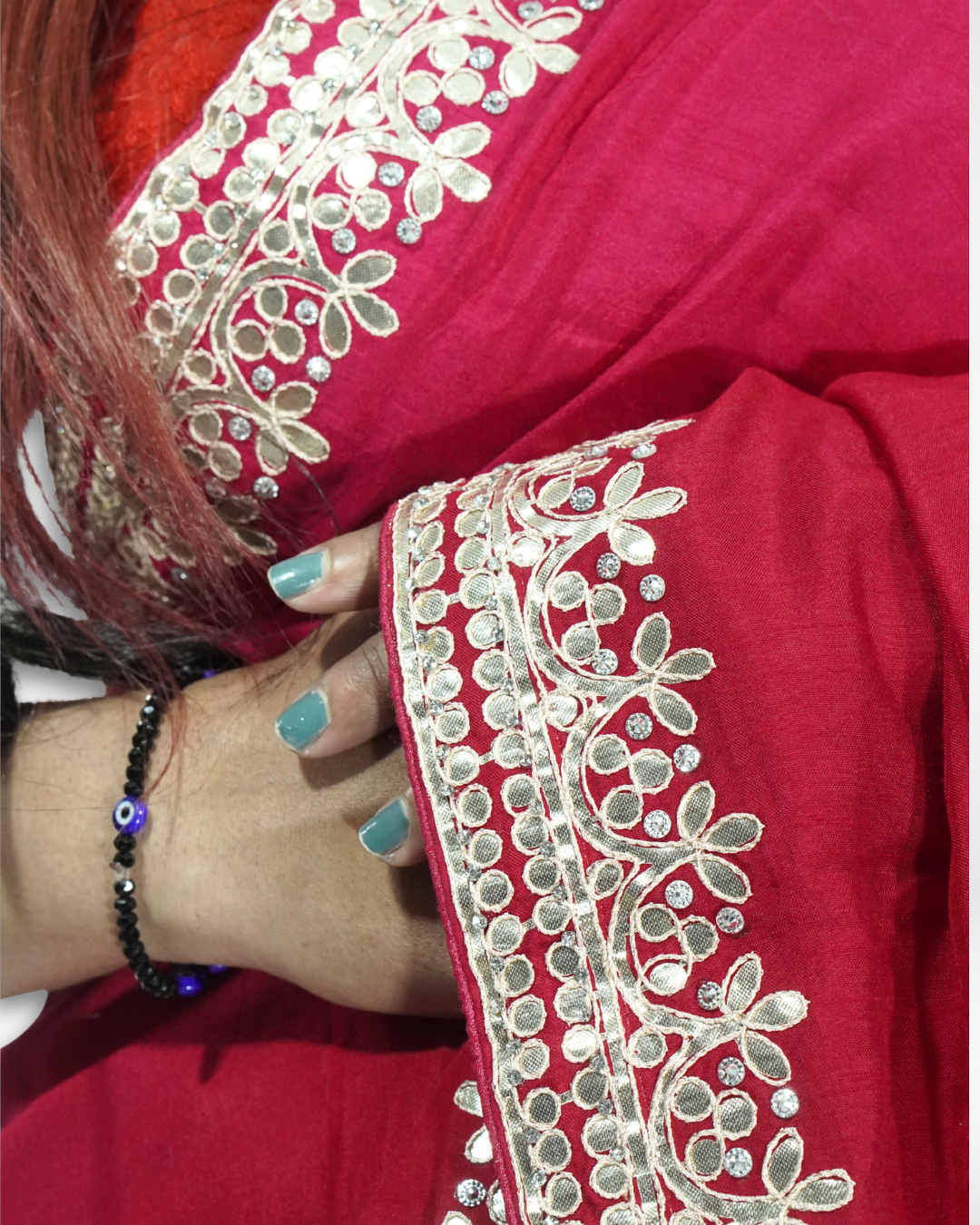 Close-up of a red saree showing intricate gold floral embroidery and stone detailing along the border, with a hand gently holding the fabric to highlight the craftsmanship and design. ✨