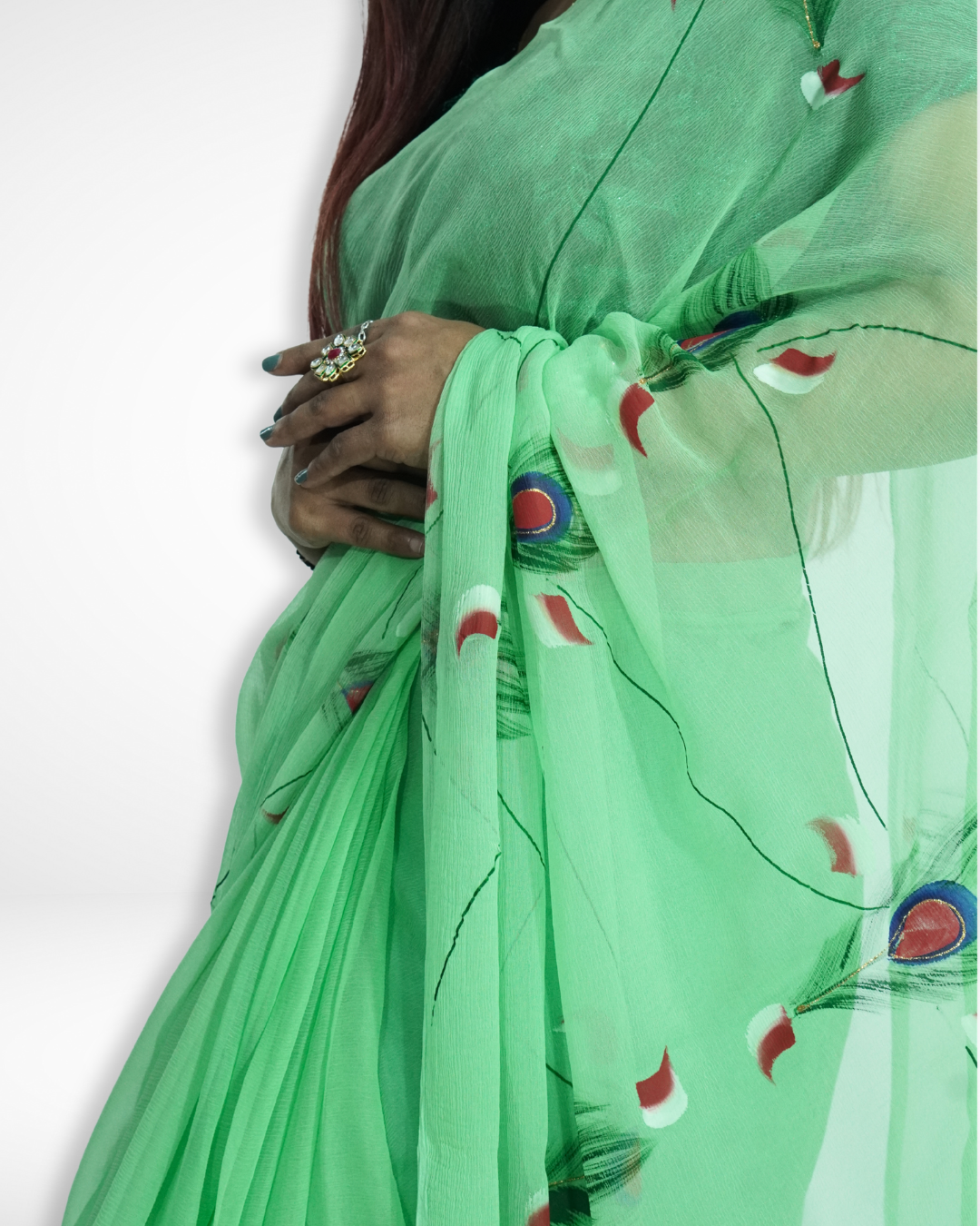 Detailed close-up of a mint green chiffon saree featuring artistic peacock feather prints and soft pleats, worn by a model with elegant jewelry, showcasing Amai Jaipur’s lightweight designer saree perfect for festive and occasion wear. ✨
