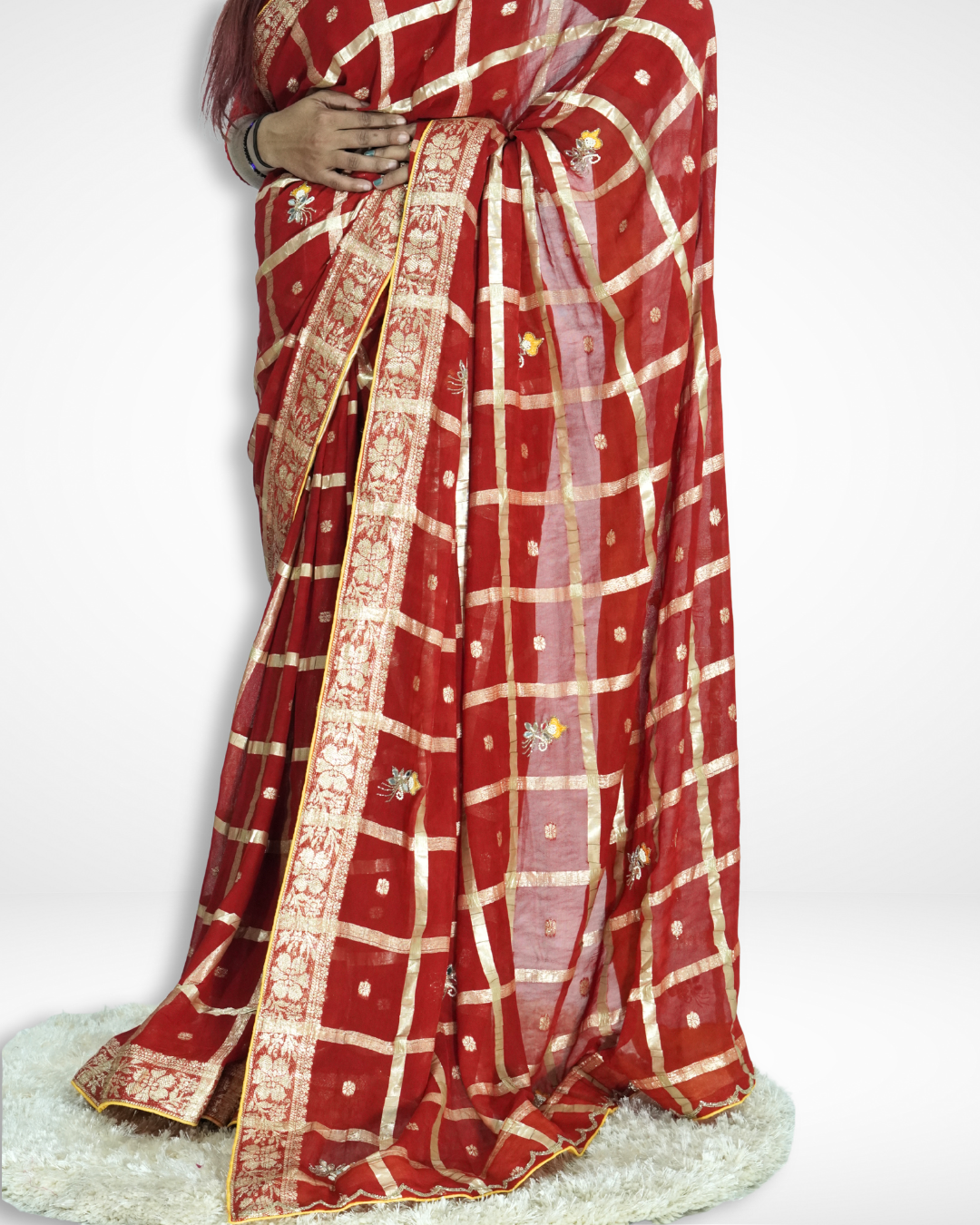 Detailed view of a red saree with shimmering gold checkered zari pattern, intricate floral zari border, and delicate embroidered motifs, elegantly draped to highlight its rich texture and festive appeal. ✨