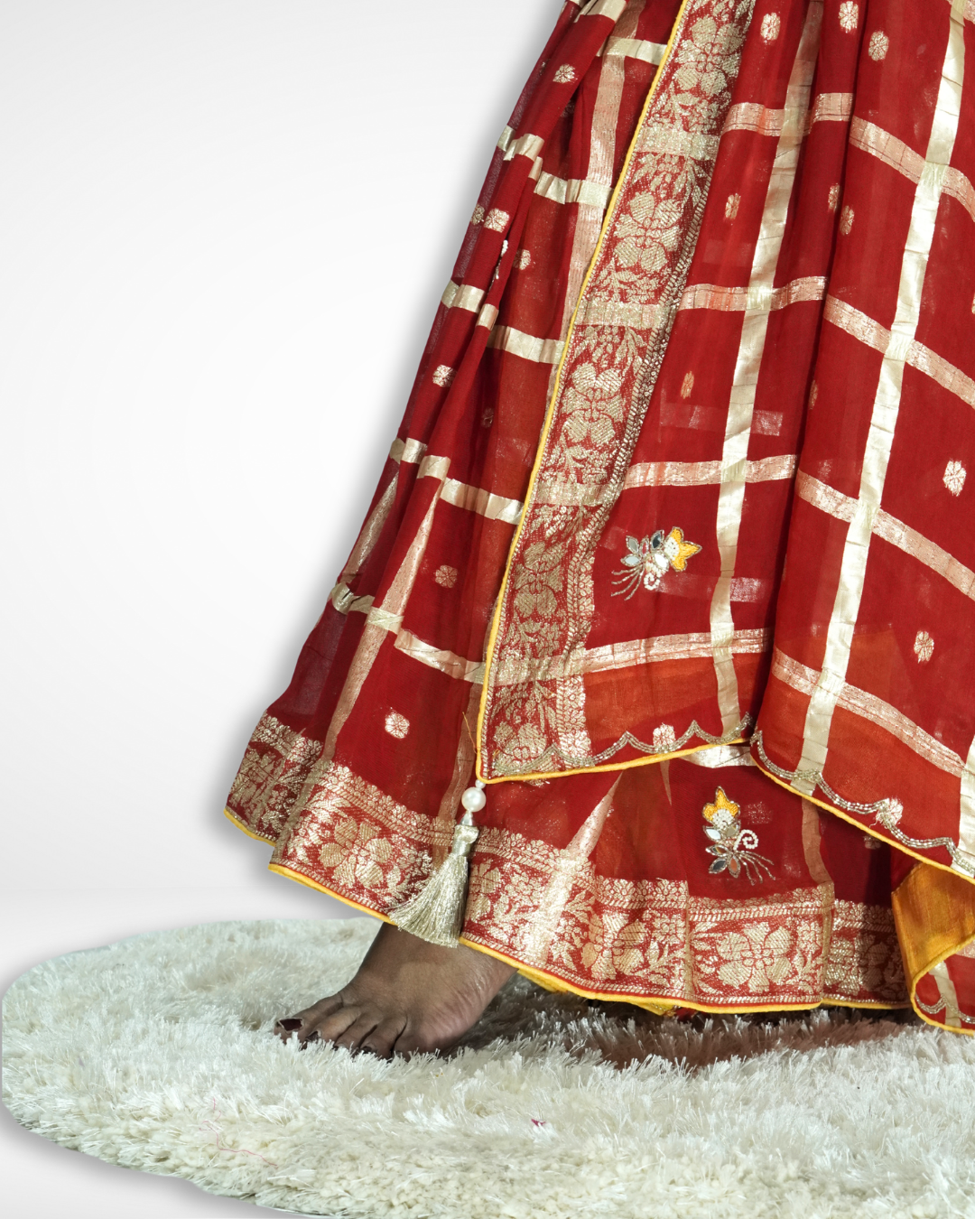 Close-up of the lower drape of a red saree featuring a shimmering gold checkered zari pattern, ornate floral zari border, and delicate embroidered motifs, finished with a decorative tassel for an elegant festive look. ✨