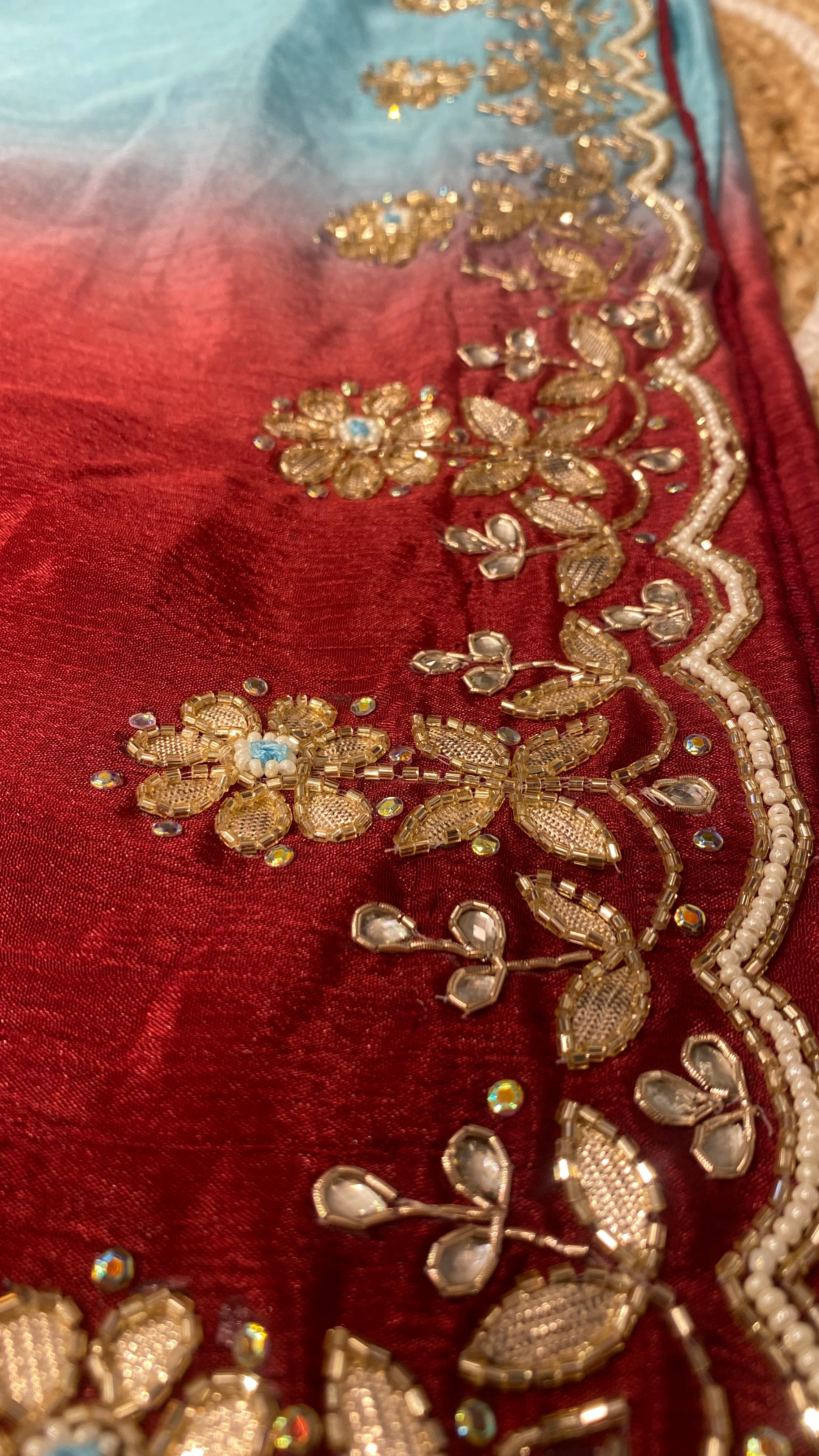 Detailed close-up of an ombre saree blending from sky blue to deep red, showcasing intricate gold floral embroidery with beadwork, crystal accents, and a scalloped pearl border along the edge. ✨
