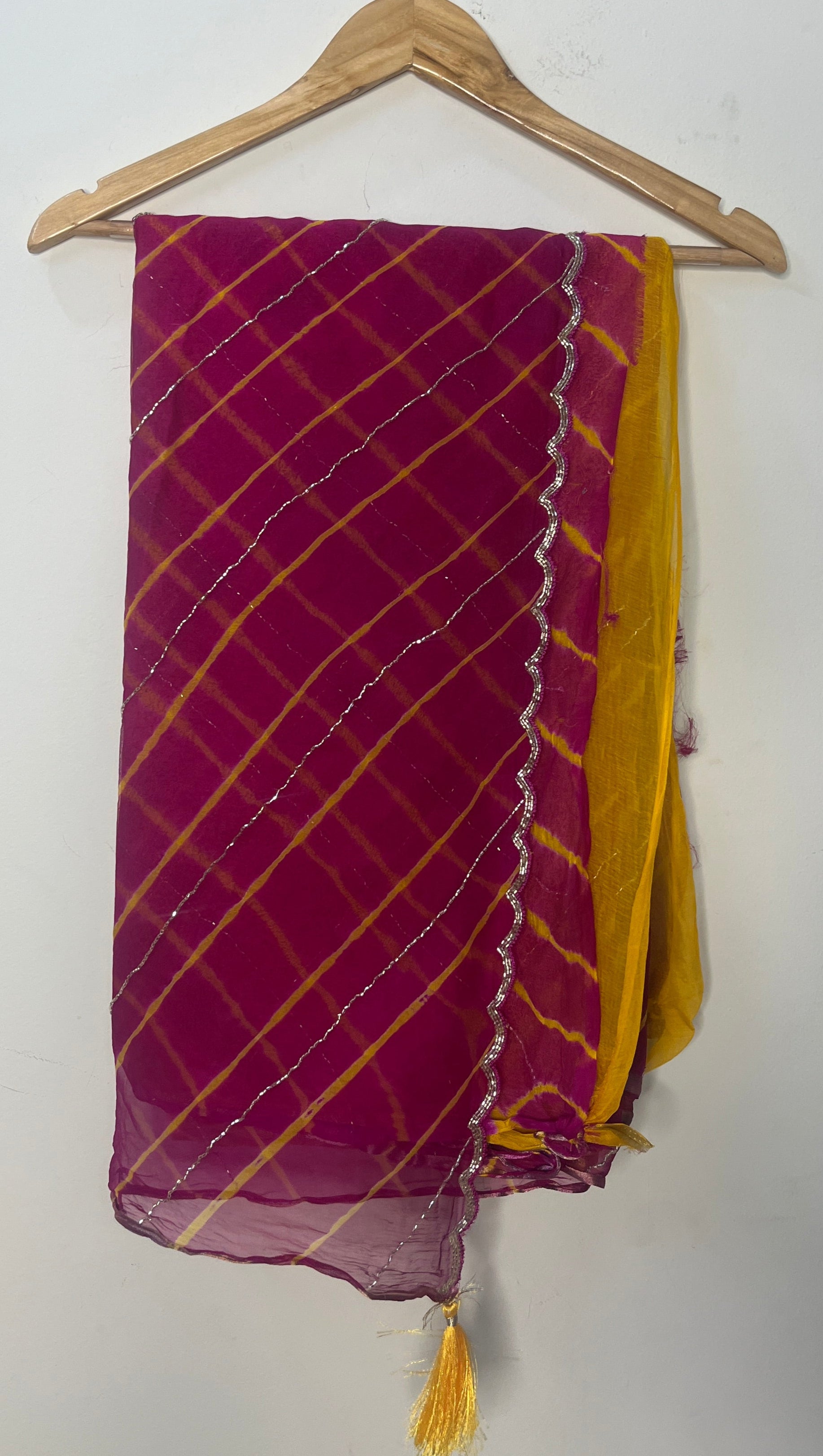 Magenta and pink tie-dye saree with diagonal yellow stripes and delicate silver shimmer lines, finished with a scalloped metallic border and mustard yellow pallu, displayed on a wooden hanger. ✨