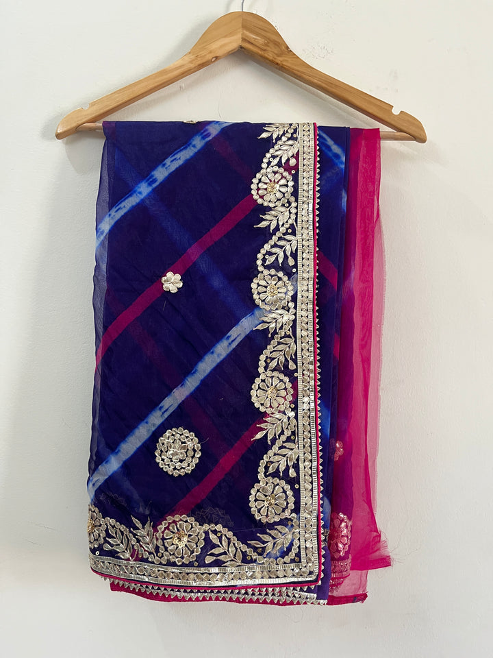 Folded navy blue and magenta saree displayed on a wooden hanger, featuring diagonal tie-dye style patterns with intricate silver floral embroidery and an ornate embellished border. ✨