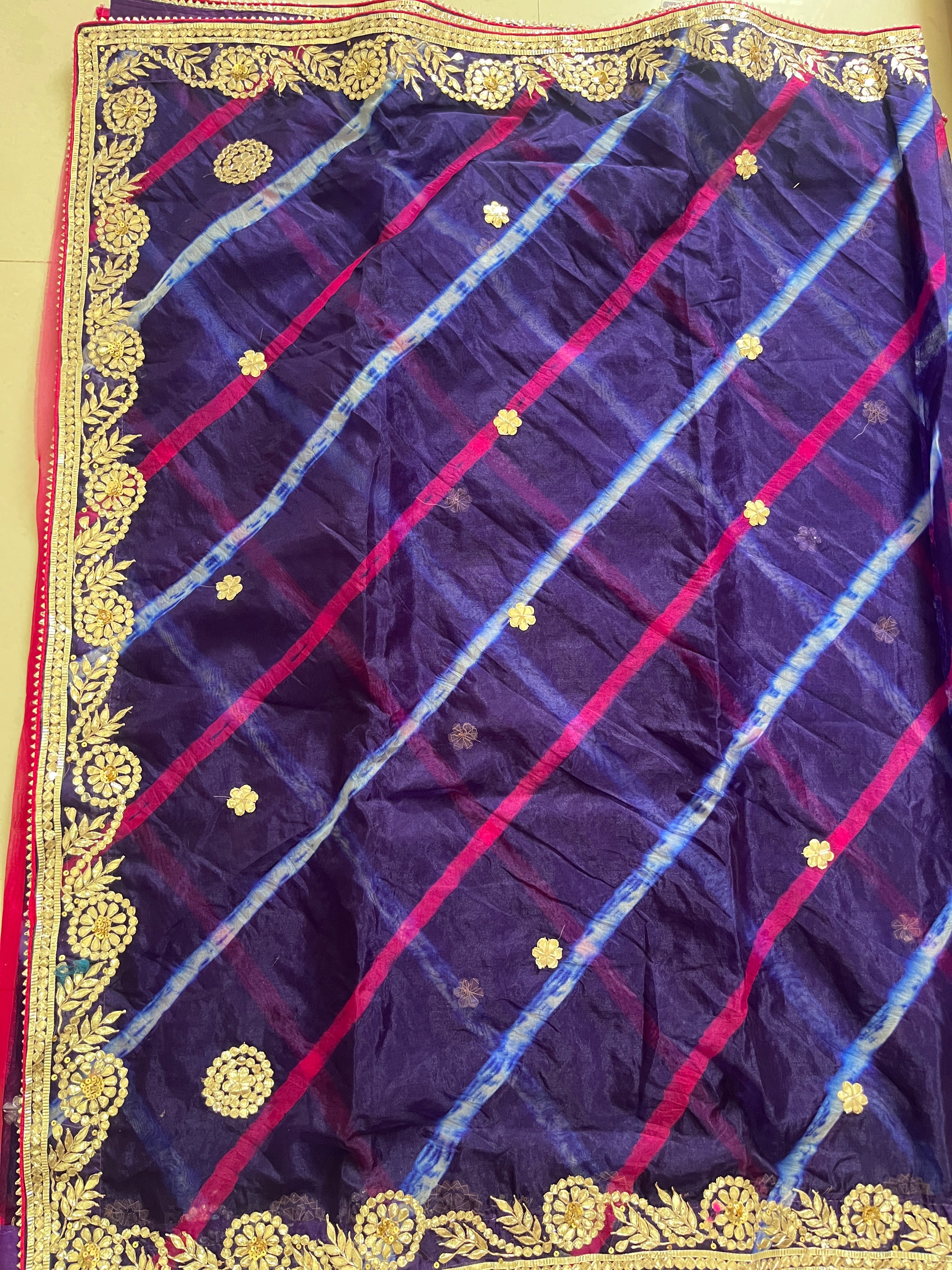 Flat lay of a deep purple saree featuring diagonal pink and sky-blue tie-dye style stripes, scattered small gold floral butta motifs, and an ornate gold embroidered border with floral vine patterns along the edges. ✨