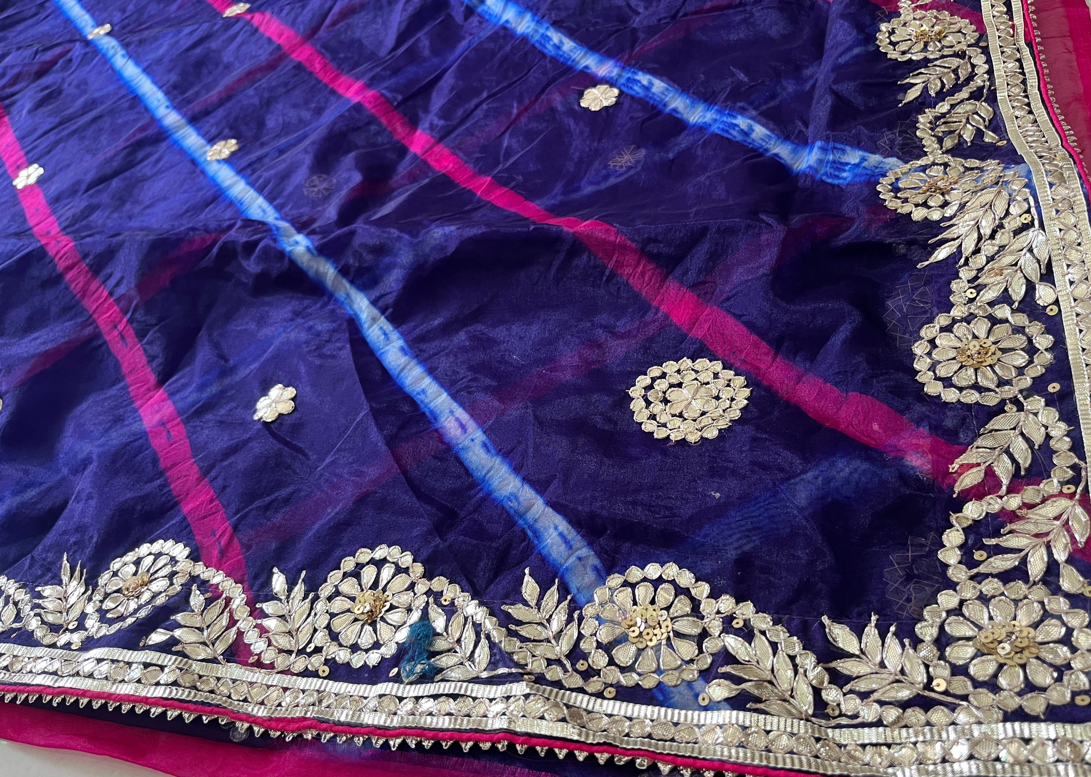 Detailed view of a deep purple saree featuring diagonal pink and sky-blue tie-dye stripes, scattered gold floral butta motifs, and a richly embroidered gold floral vine border with sequins along the edge. ✨