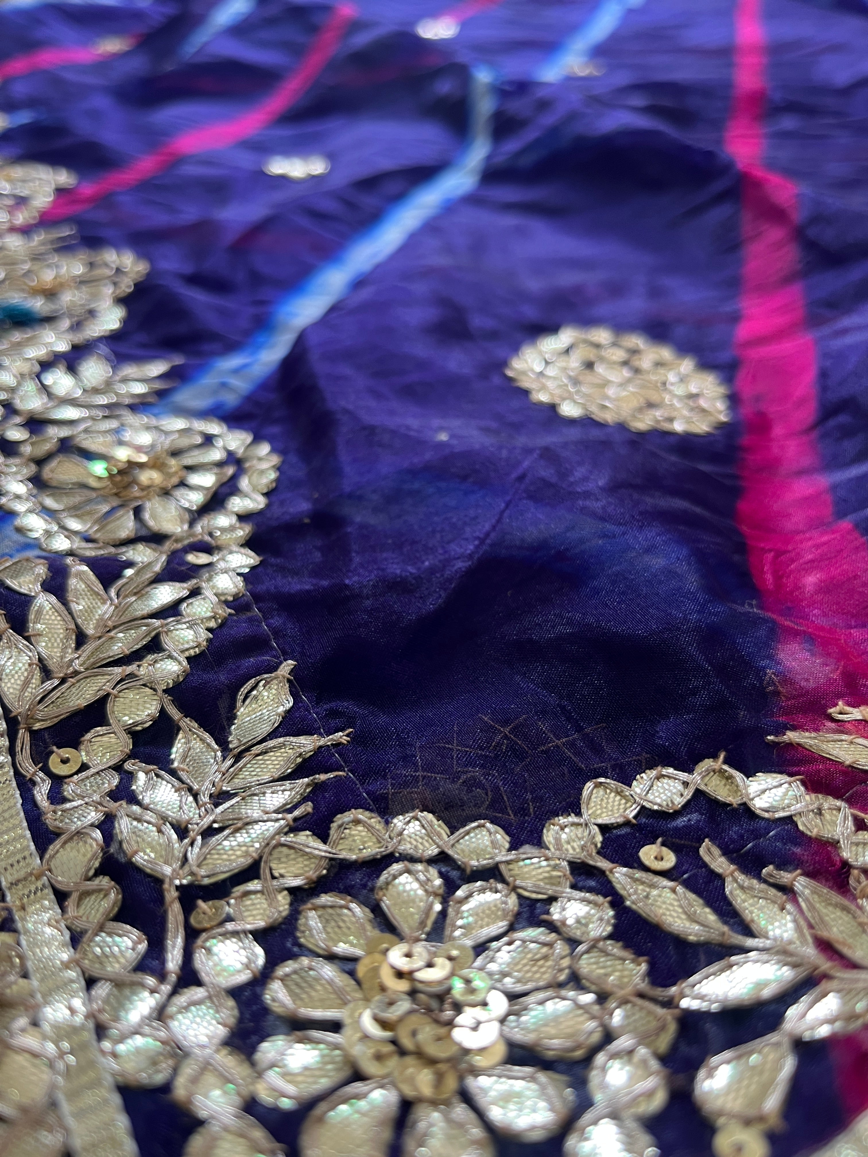 Close-up macro shot of a deep purple saree fabric showing shimmering gold floral embroidery and sequins along the ornate border, with diagonal pink and blue stripes and small gold butta motifs scattered across the textured fabric. ✨