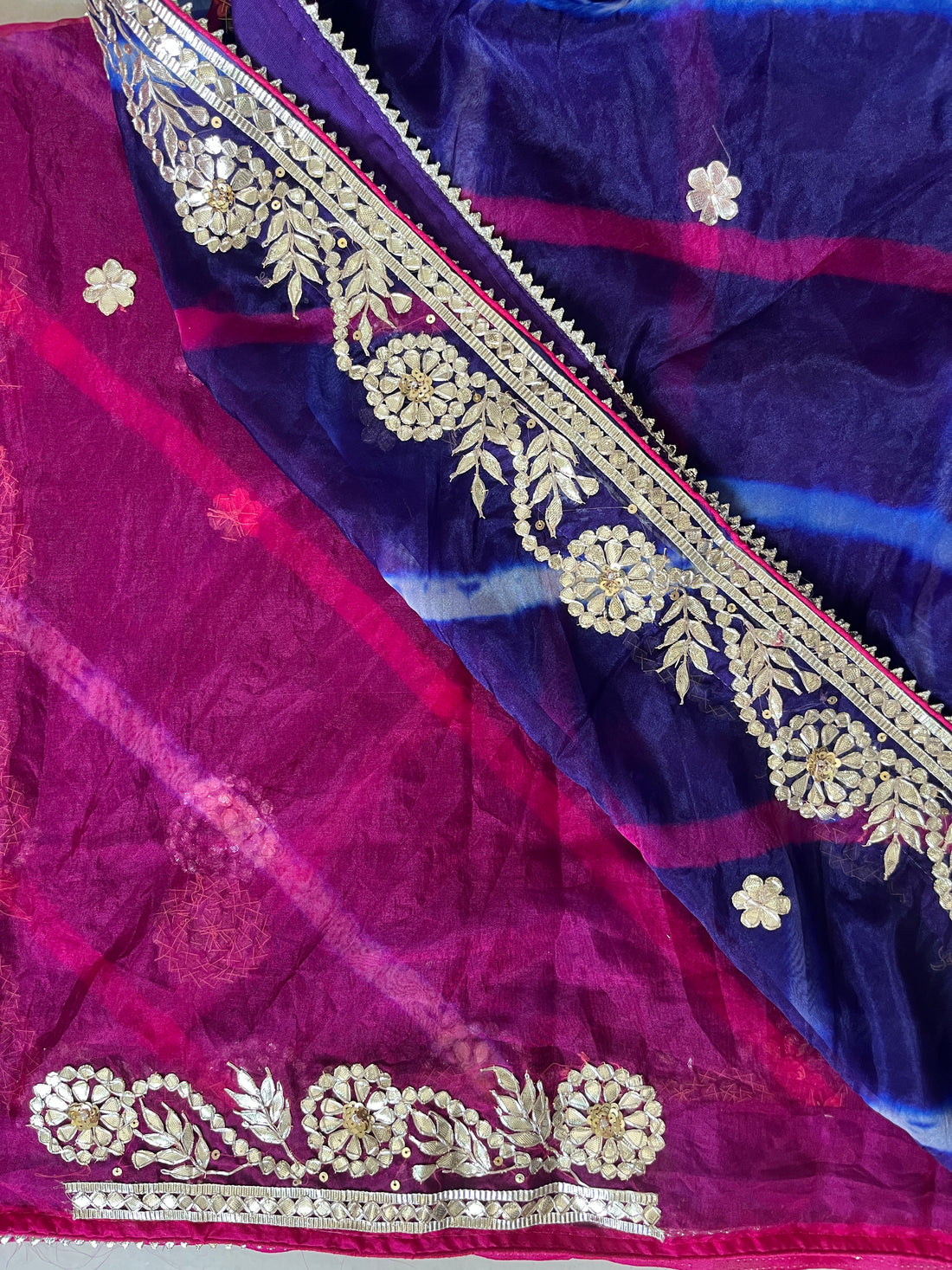 Close-up of a vibrant saree showing contrasting sections of deep purple and magenta with diagonal blue and pink tie-dye stripes, decorated with small gold floral butta motifs and an ornate gold embroidered border with floral vine detailing and sequins. ✨
