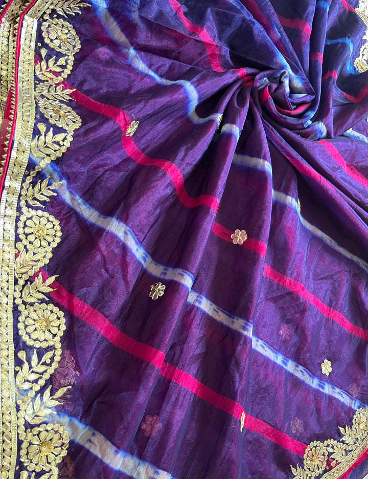 Close-up of a rich purple saree arranged in soft folds, featuring pink and blue tie-dye style stripes, delicate gold floral butta motifs, and an ornate gold embroidered border along the edge. ✨