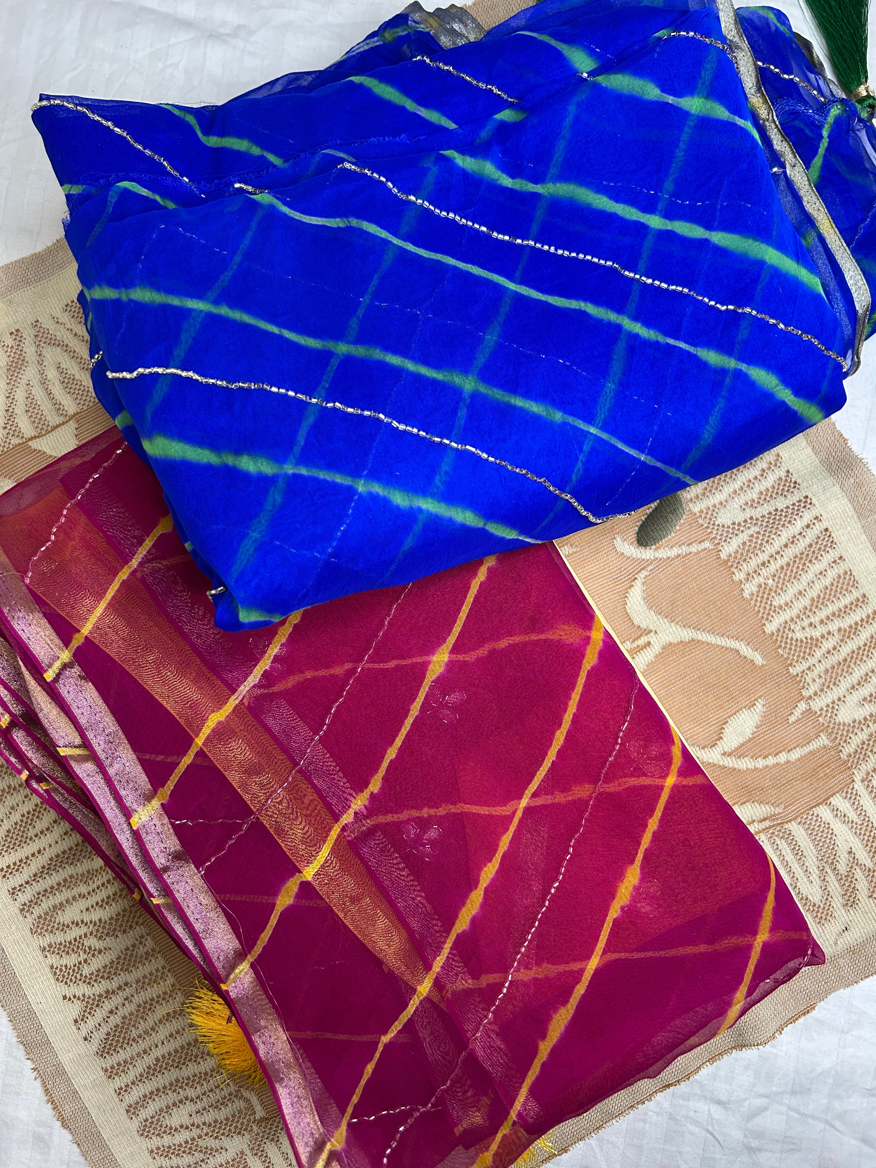 Two folded tie-dye sarees displayed together—one in royal blue with green stripes and silver shimmer lines, and the other in magenta with yellow stripes and silver accents—laid on a patterned beige fabric background. ✨