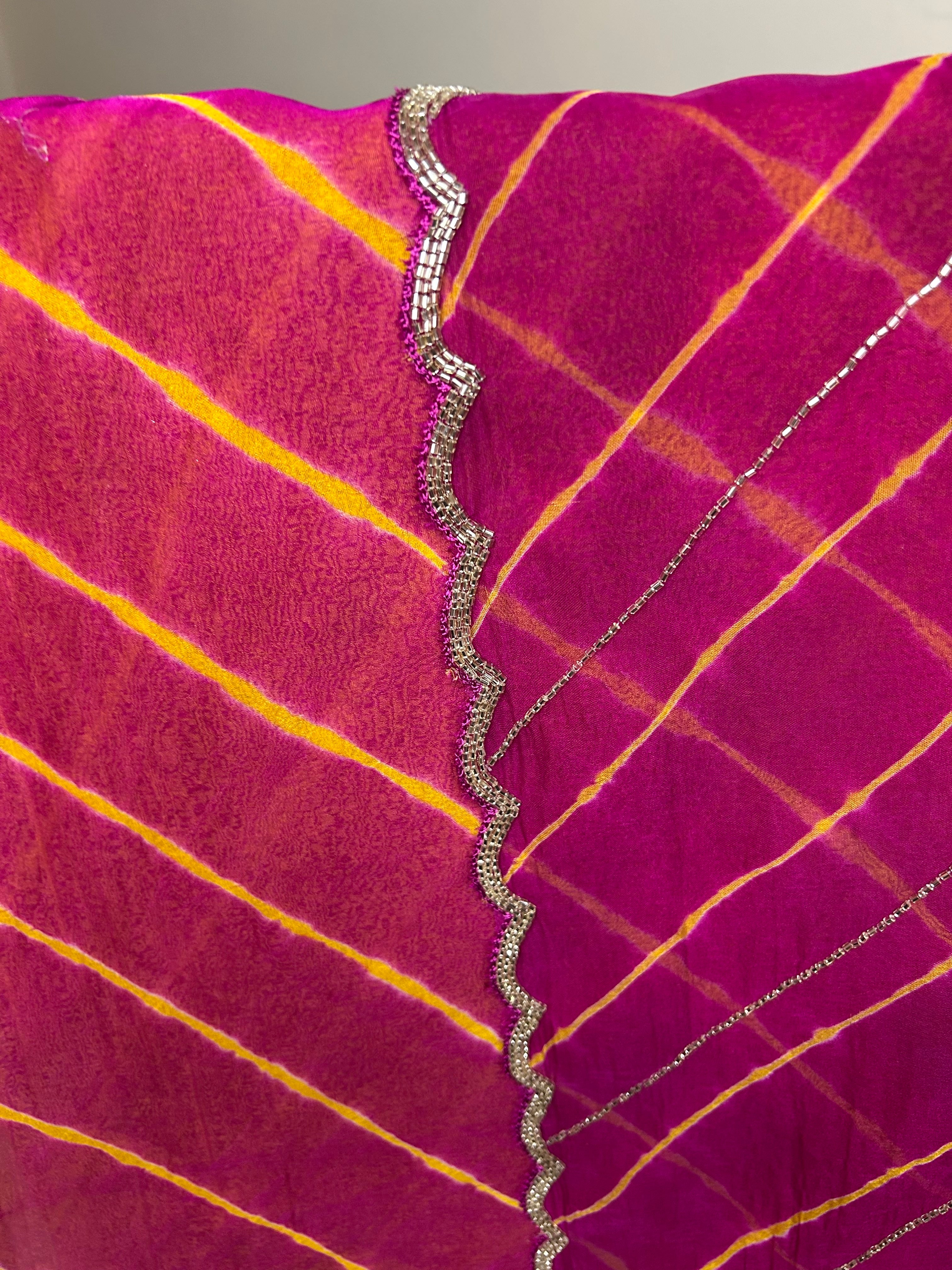 Close-up of a vibrant magenta and pink saree with traditional yellow tie-dye stripes, highlighted by delicate silver shimmer lines and a scalloped metallic border detail. ✨