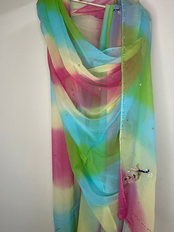 Sheer pastel saree draped on a hanger featuring a soft multicolor gradient of sky blue, lime green, pink, and cream, embellished with scattered crystal-like stones, a delicate beaded border, and a small embroidered peacock motif near the edge. ✨