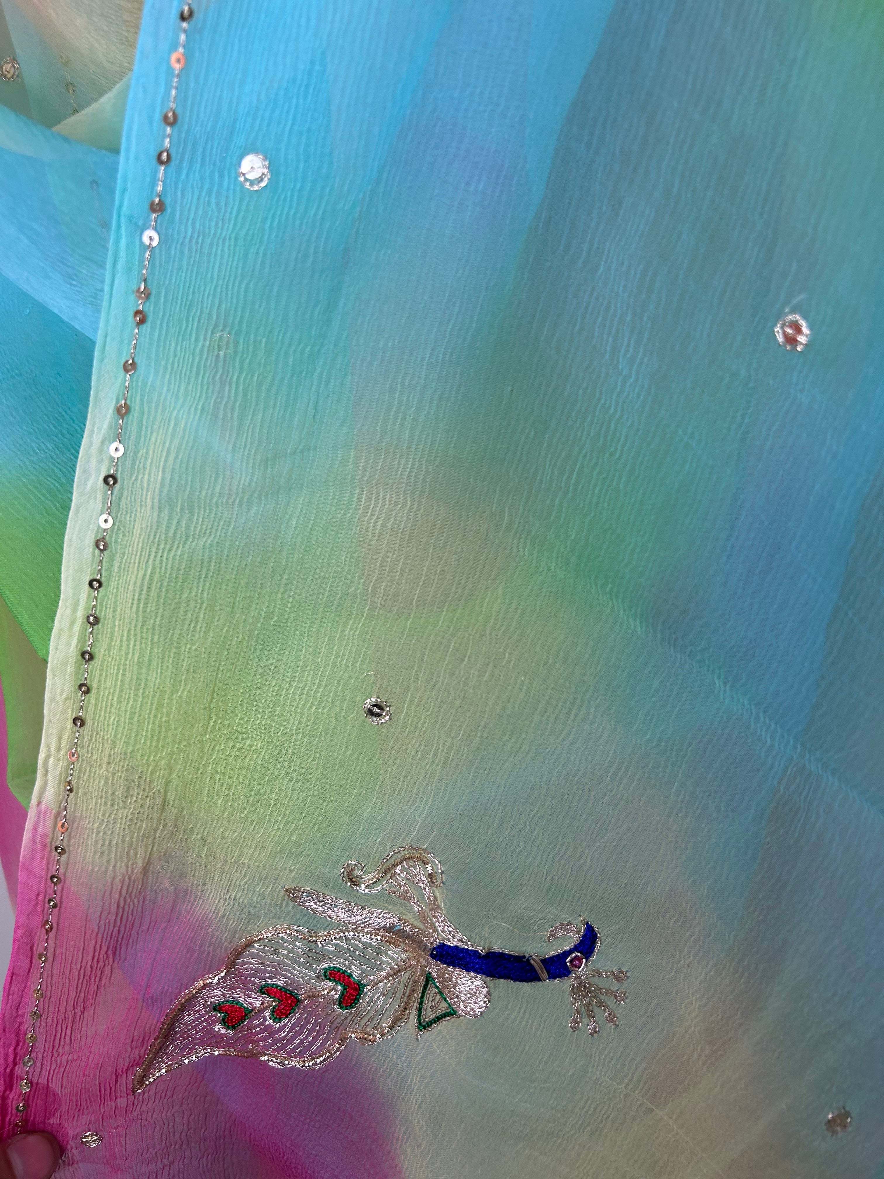 Close-up of a sheer pastel saree showing a soft gradient of sky blue, lime green, and pink fabric, embellished with small crystal-like stones, a delicate sequin border, and an embroidered peacock motif with blue, silver, and pink threadwork. ✨