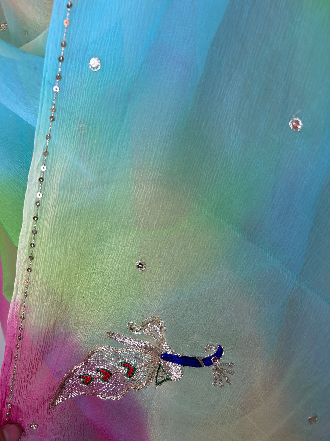 Close-up of a sheer pastel saree showing a soft gradient of sky blue, lime green, and pink fabric, embellished with small crystal-like stones, a delicate sequin border, and an embroidered peacock motif with blue, silver, and pink threadwork. ✨