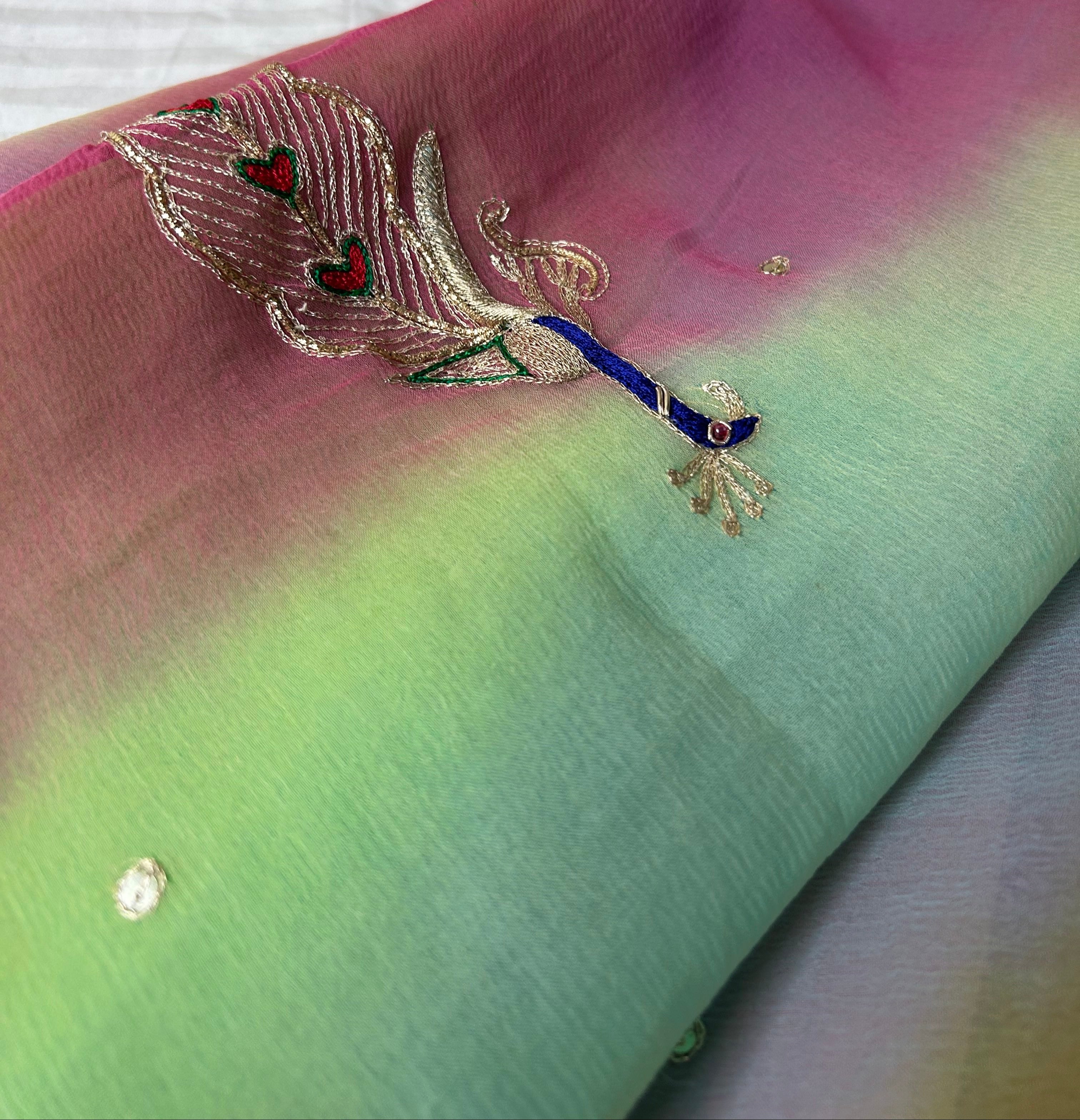 Close-up of a pastel gradient saree folded neatly, featuring soft shades of pink, mint green, and sky blue, embellished with small crystal-like stones and an embroidered peacock motif with blue, silver, and red threadwork. ✨