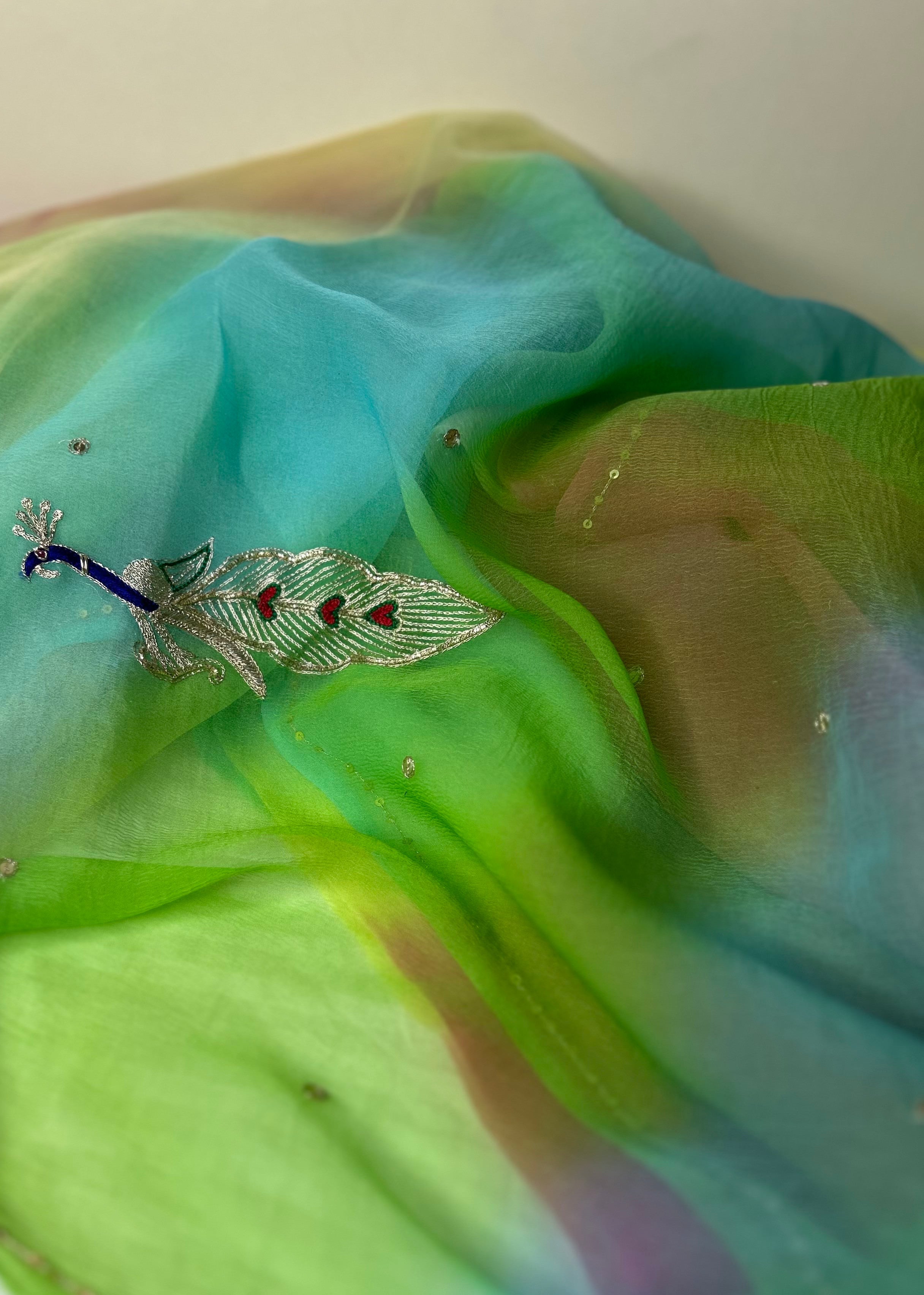 Close-up of a sheer pastel gradient saree in flowing folds, blending aqua blue, lime green, and soft peach tones, adorned with tiny crystal-like embellishments and an embroidered peacock motif with blue, silver, and red detailing. ✨