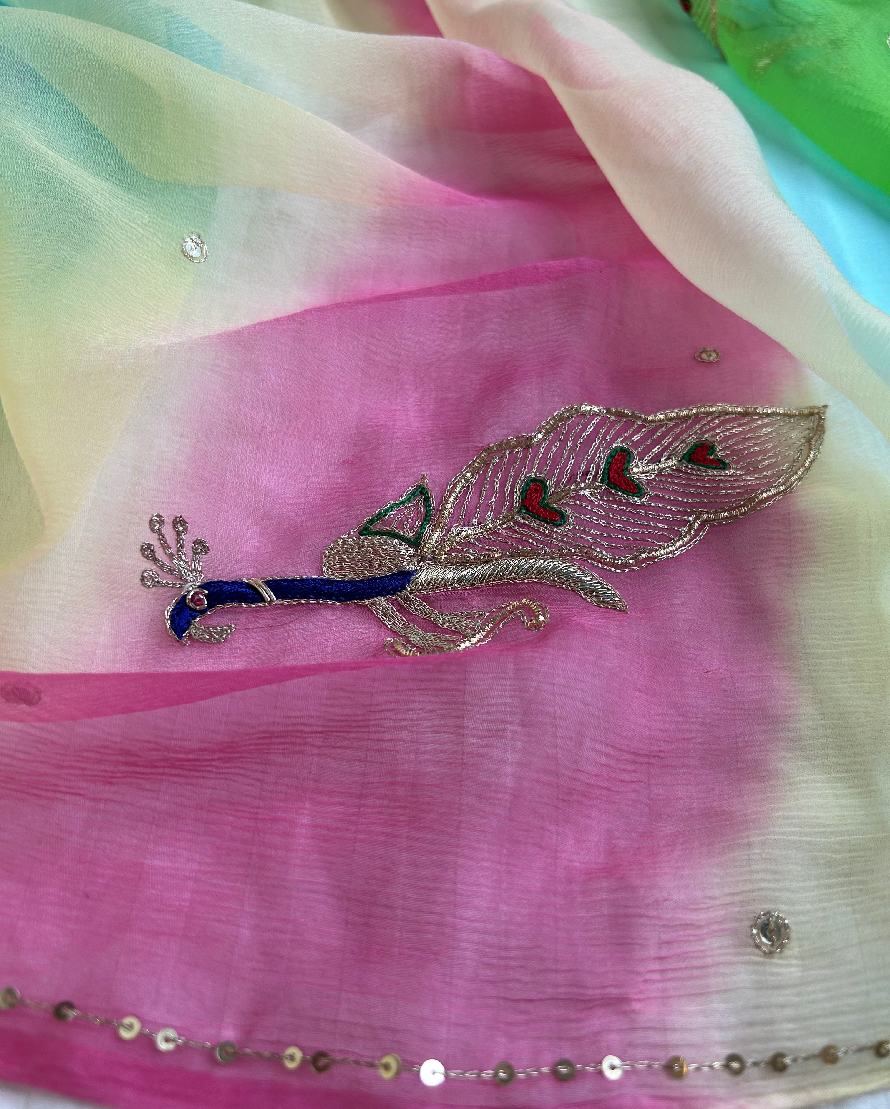 Close-up of a sheer pastel saree with soft pink, cream, and light blue gradient tones, featuring delicate crystal-like embellishments, a fine sequin border, and an embroidered peacock motif with blue, silver, and red detailing. ✨