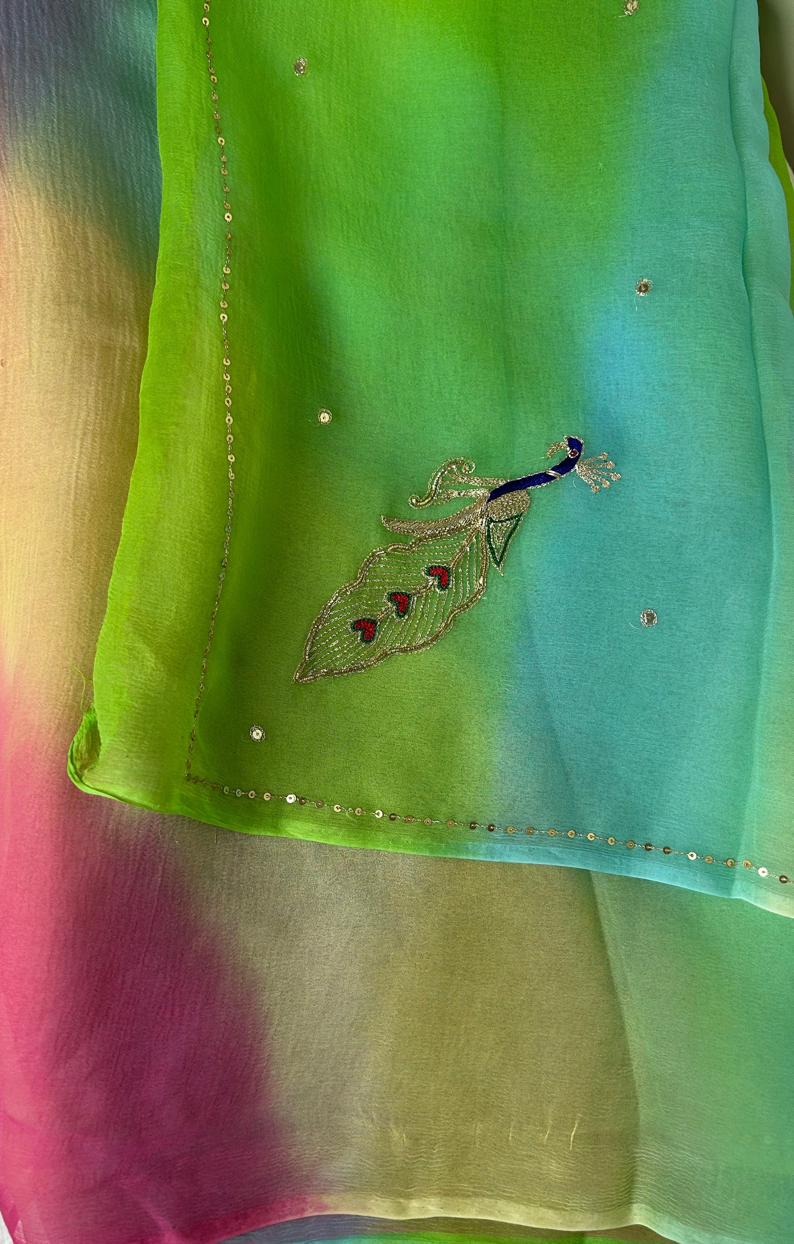 Sheer pastel gradient saree featuring vibrant shades of lime green, aqua blue, soft pink, and cream, adorned with scattered crystal-like embellishments, a delicate sequin border, and an embroidered peacock motif with blue, silver, and red detailing. ✨