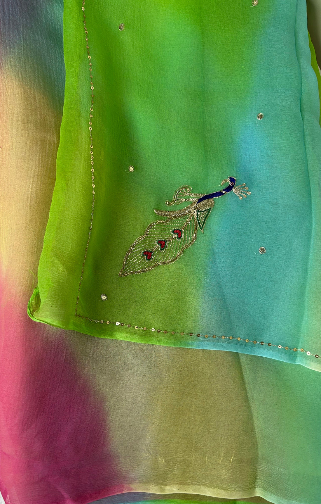 Sheer pastel gradient saree featuring vibrant shades of lime green, aqua blue, soft pink, and cream, adorned with scattered crystal-like embellishments, a delicate sequin border, and an embroidered peacock motif with blue, silver, and red detailing. ✨