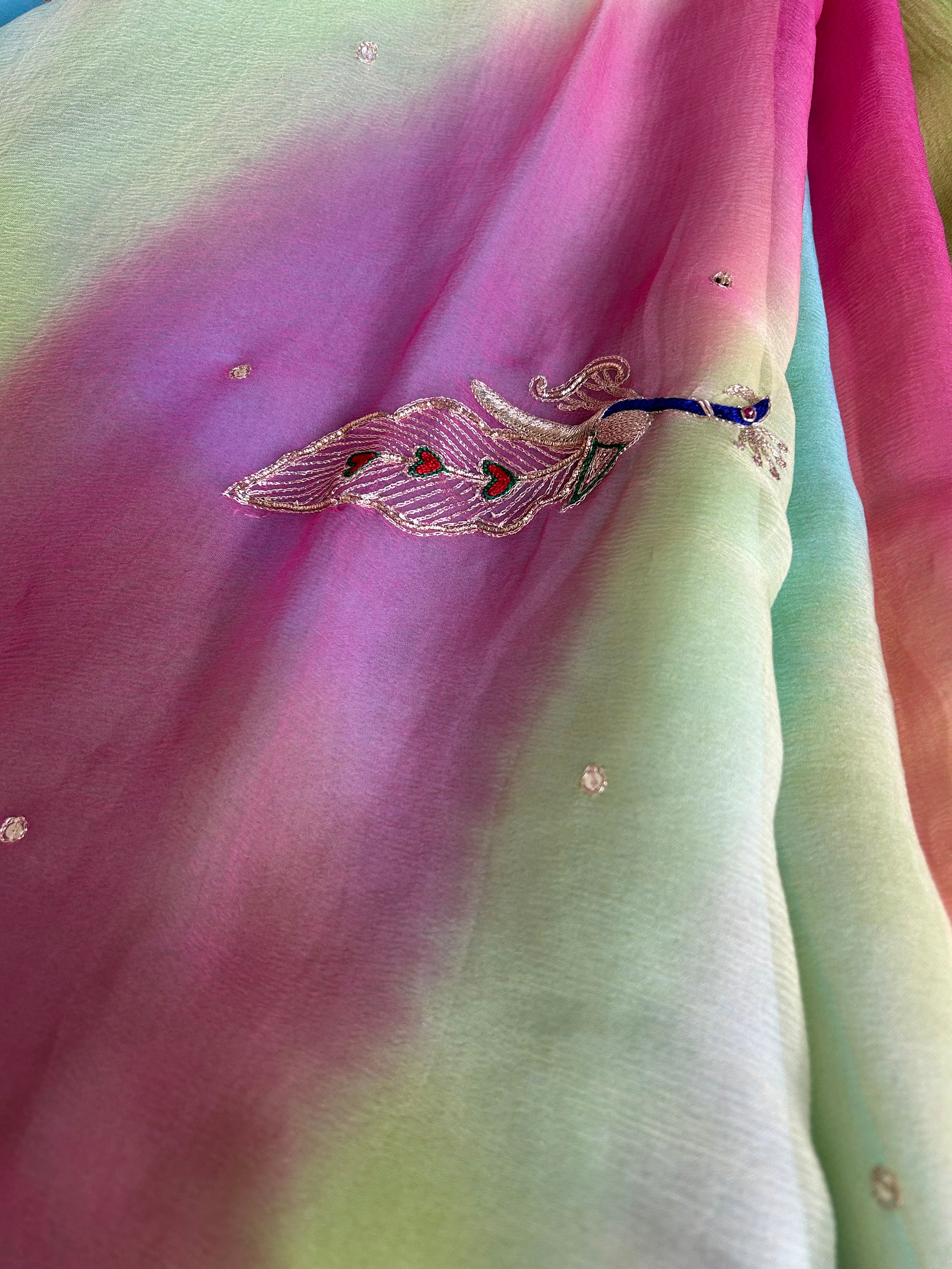 Close-up of a sheer pastel gradient saree blending pink, mint green, and soft blue tones, decorated with small crystal-like embellishments and an embroidered peacock motif featuring blue, silver, and red threadwork. ✨