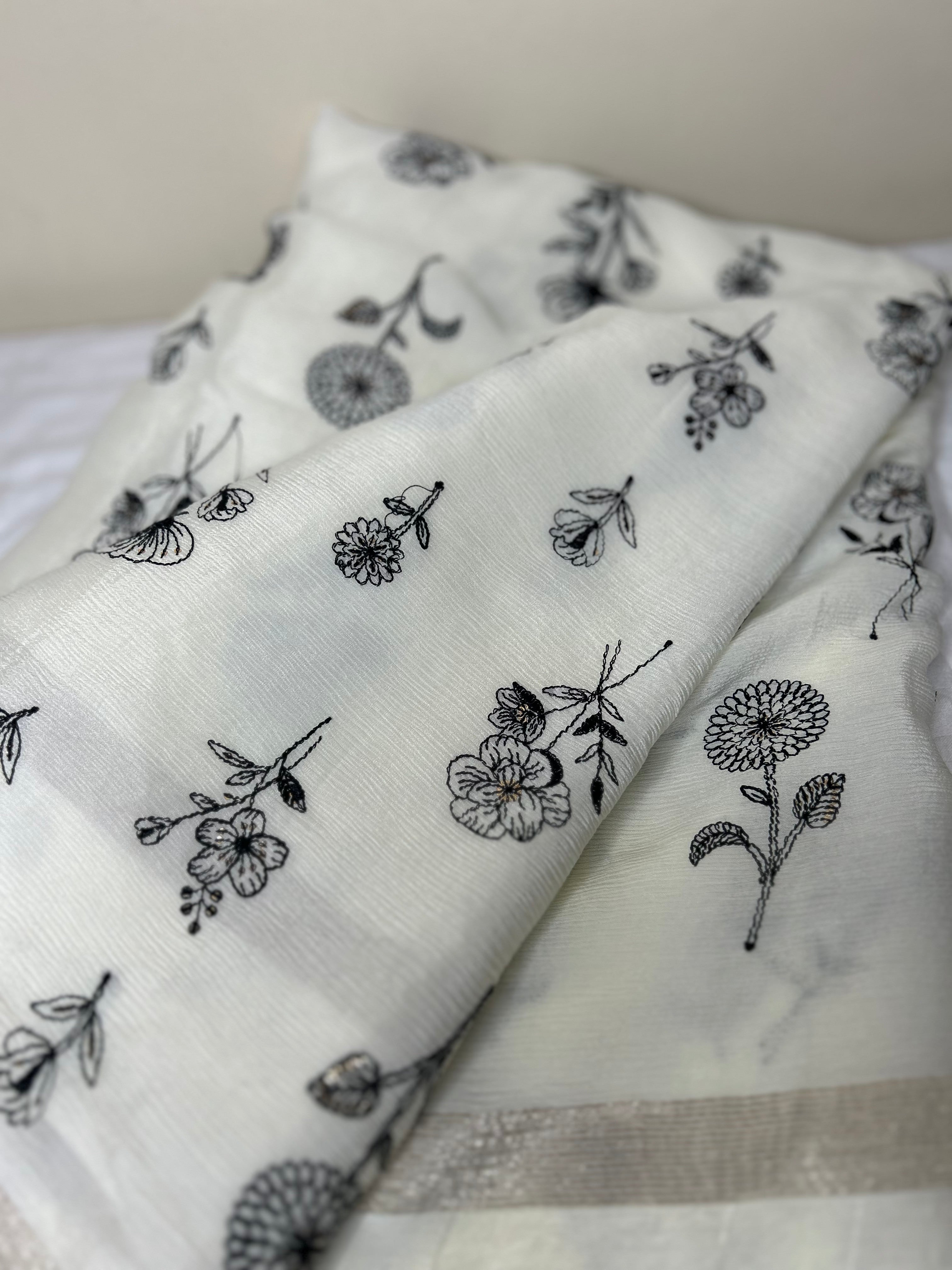 Close-up of an elegant off-white saree fabric featuring delicate black floral thread embroidery scattered across the soft textured material, with a subtle silver border visible along the edge. ✨