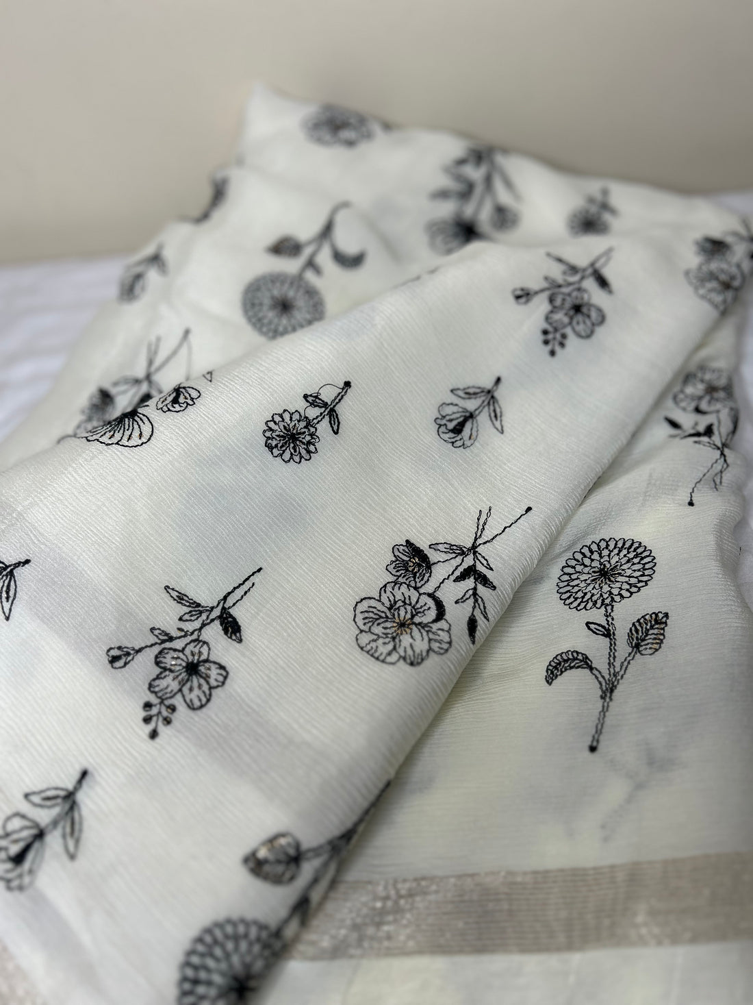 Close-up of an elegant off-white saree fabric featuring delicate black floral thread embroidery scattered across the soft textured material, with a subtle silver border visible along the edge. ✨