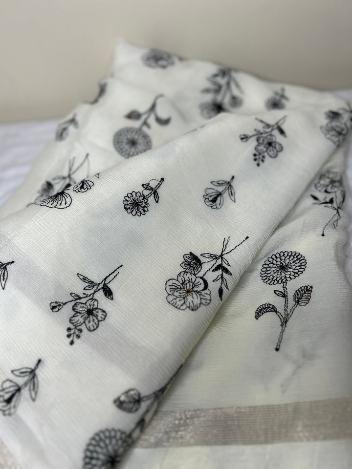 Close-up of an elegant off-white saree fabric featuring delicate black floral thread embroidery scattered across the soft textured material, with a subtle silver border visible along the edge. ✨