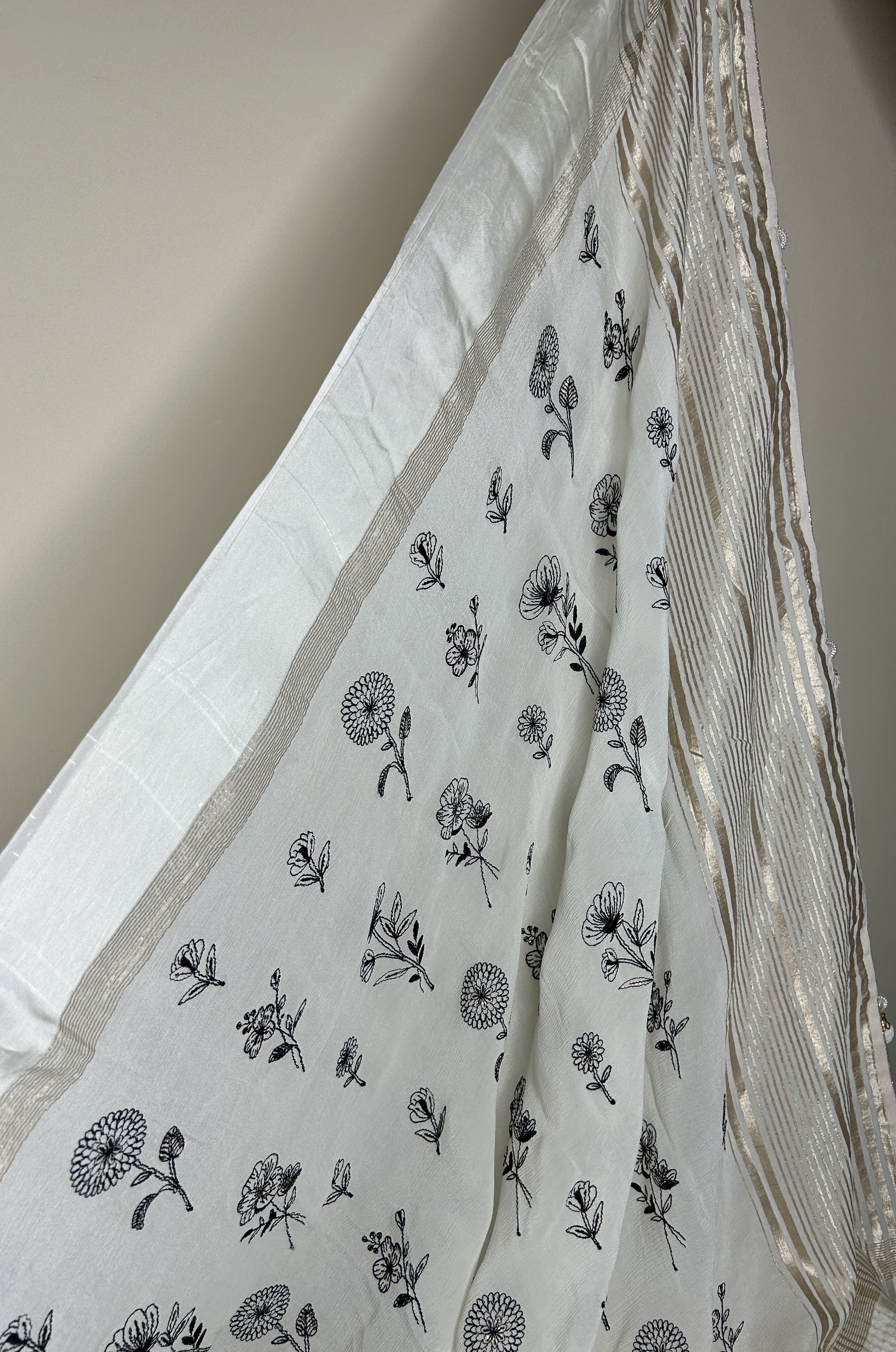 Elegant white saree with delicate black floral prints scattered across the fabric, finished with a subtle gold striped border that adds a refined, minimalistic look. ✨