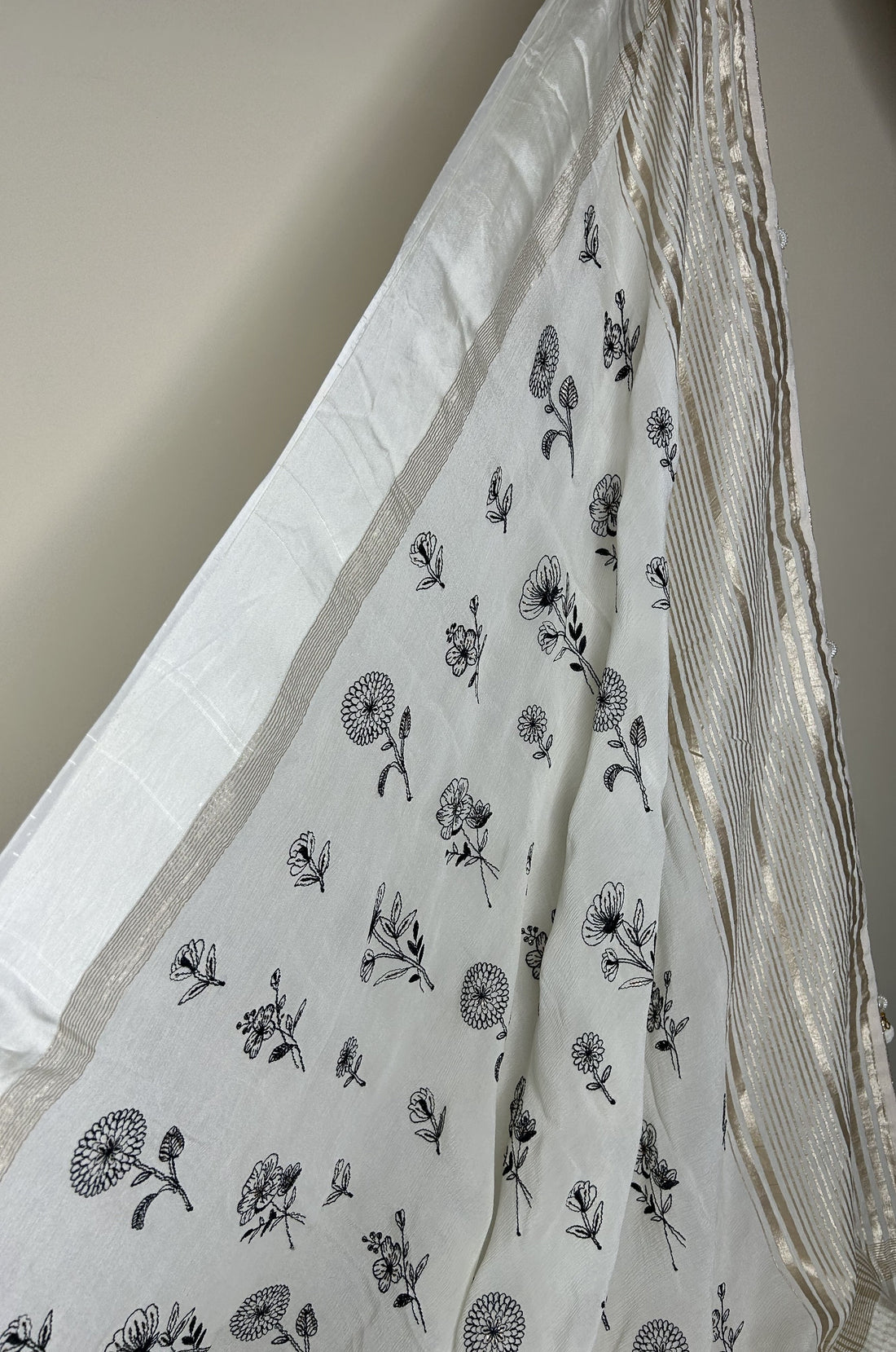 Elegant white saree with delicate black floral prints scattered across the fabric, finished with a subtle gold striped border that adds a refined, minimalistic look. ✨