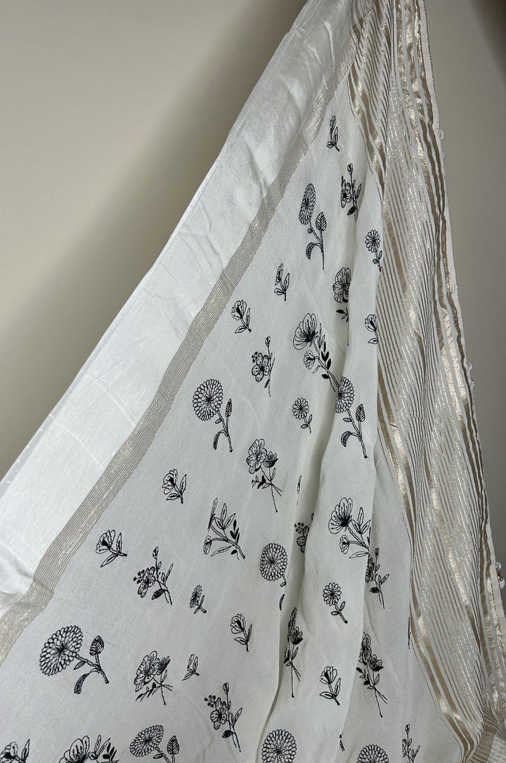 Elegant white saree with delicate black floral prints scattered across the fabric, finished with a subtle gold striped border that adds a refined, minimalistic look. ✨