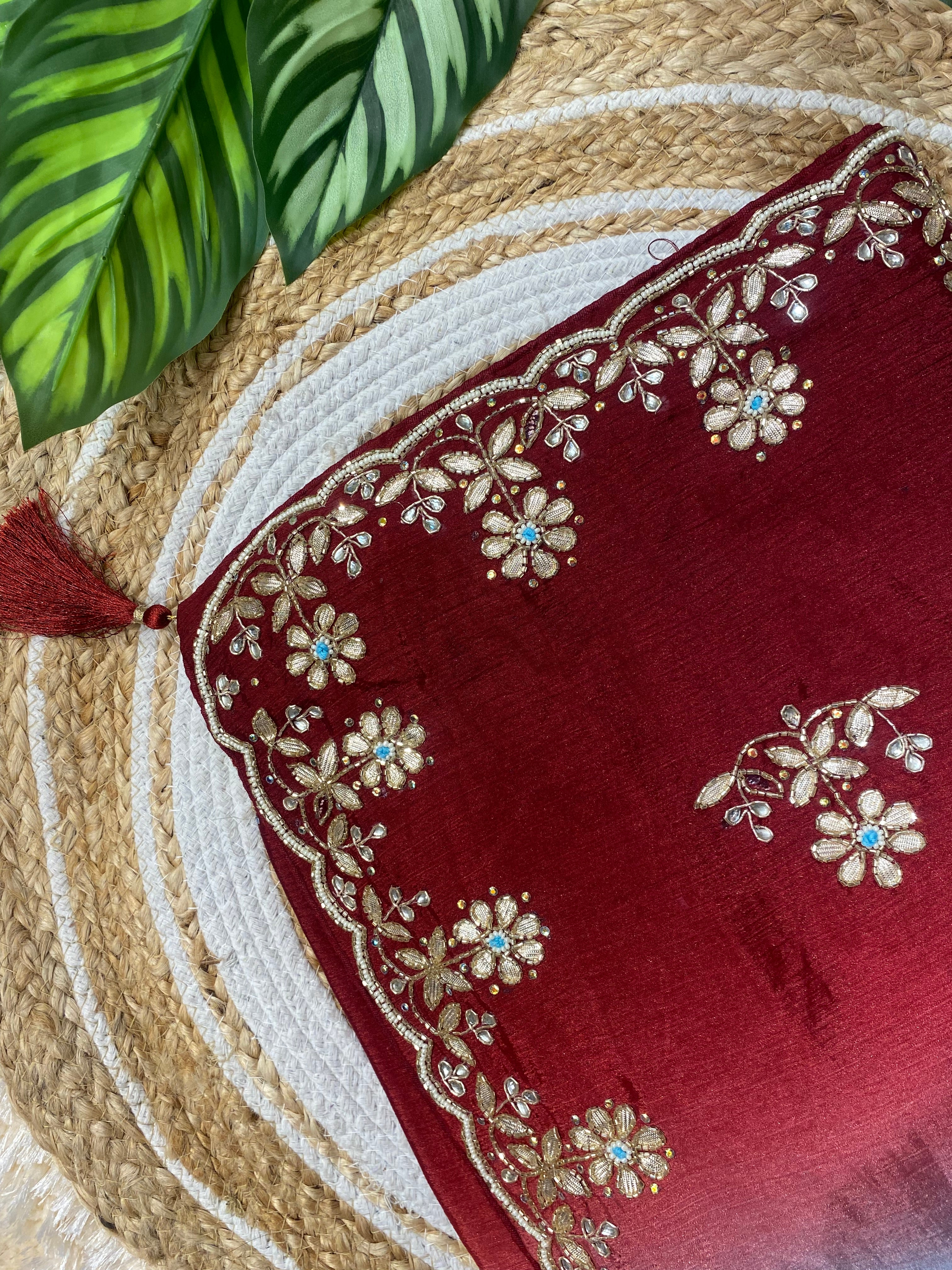 Close-up of a rich red saree corner featuring intricate gold floral embroidery along a scalloped border, accented with small blue stone details and a decorative tassel, displayed on a woven jute mat with green leaves in the background. ✨