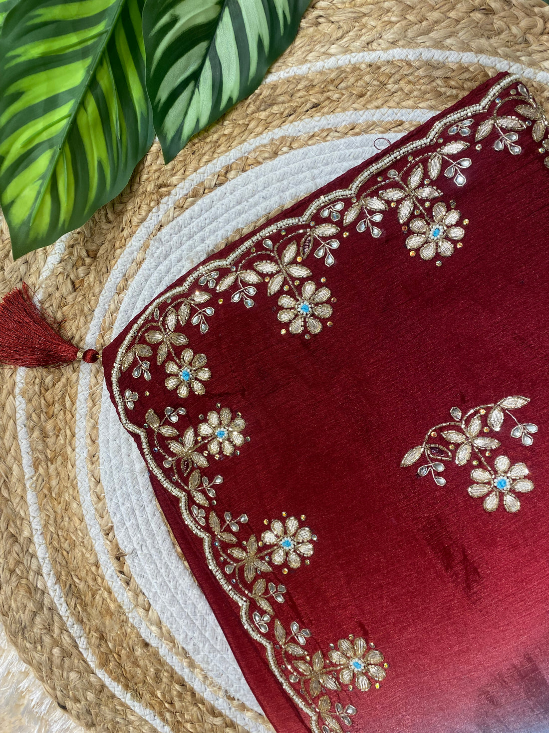 Close-up of a rich red saree corner featuring intricate gold floral embroidery along a scalloped border, accented with small blue stone details and a decorative tassel, displayed on a woven jute mat with green leaves in the background. ✨