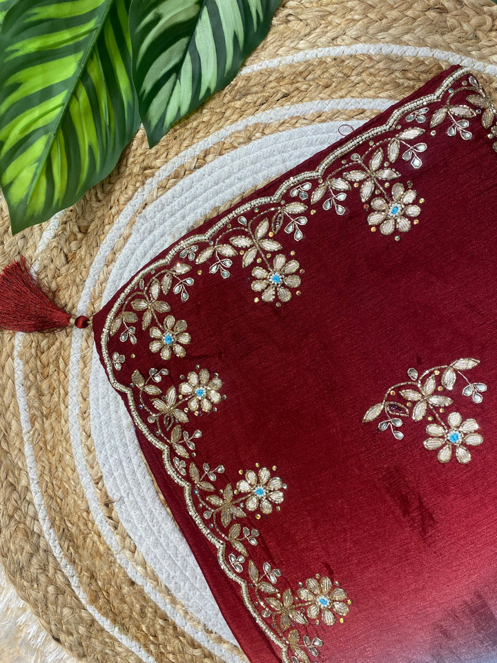 Close-up of a rich red saree corner featuring intricate gold floral embroidery along a scalloped border, accented with small blue stone details and a decorative tassel, displayed on a woven jute mat with green leaves in the background. ✨