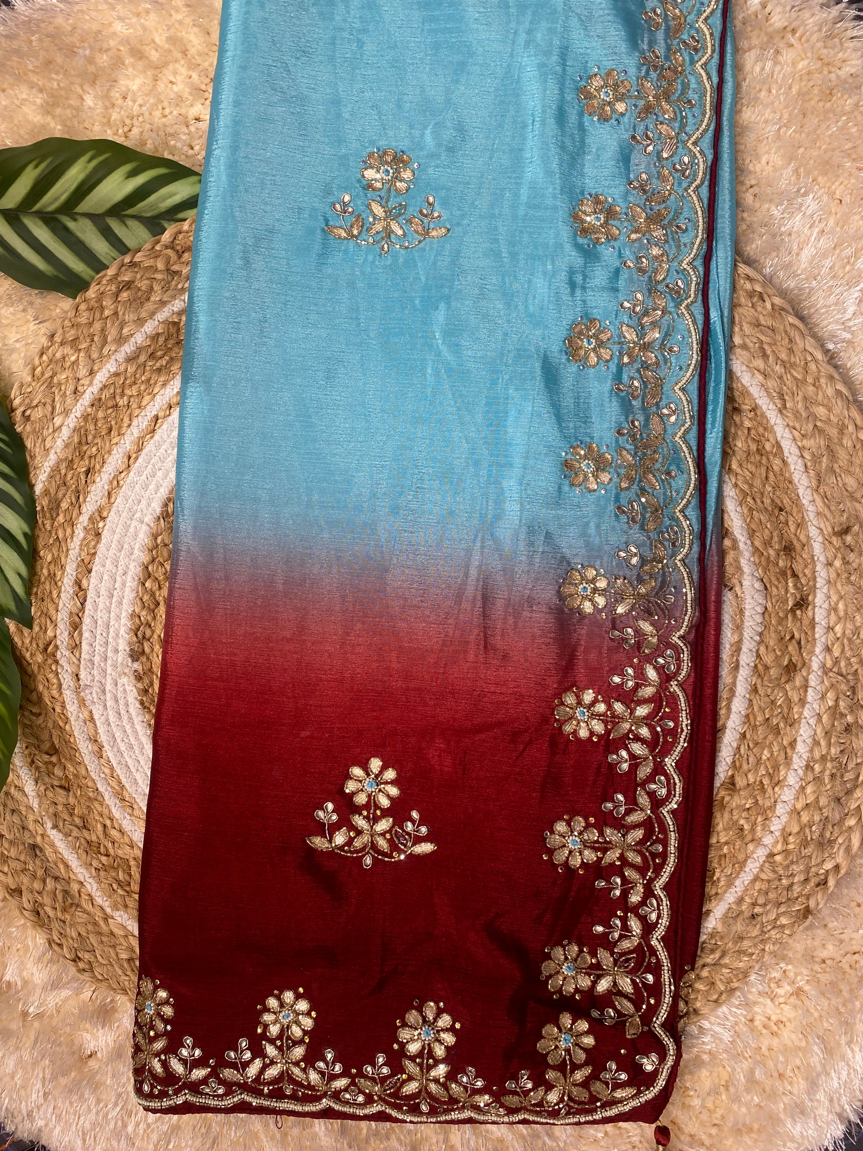 Folded ombre saree transitioning from sky blue to deep red, adorned with delicate gold floral embroidery and a scalloped border, styled on a woven jute mat with decorative green leaves and a soft textured backdrop. ✨