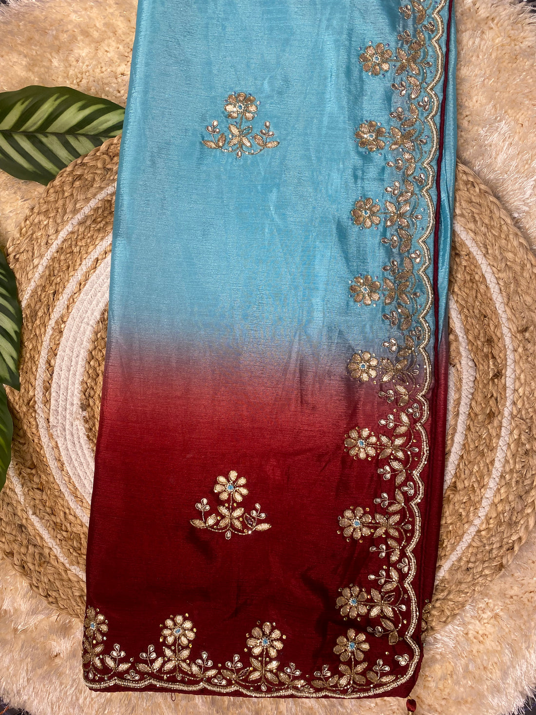 Folded ombre saree transitioning from sky blue to deep red, adorned with delicate gold floral embroidery and a scalloped border, styled on a woven jute mat with decorative green leaves and a soft textured backdrop. ✨