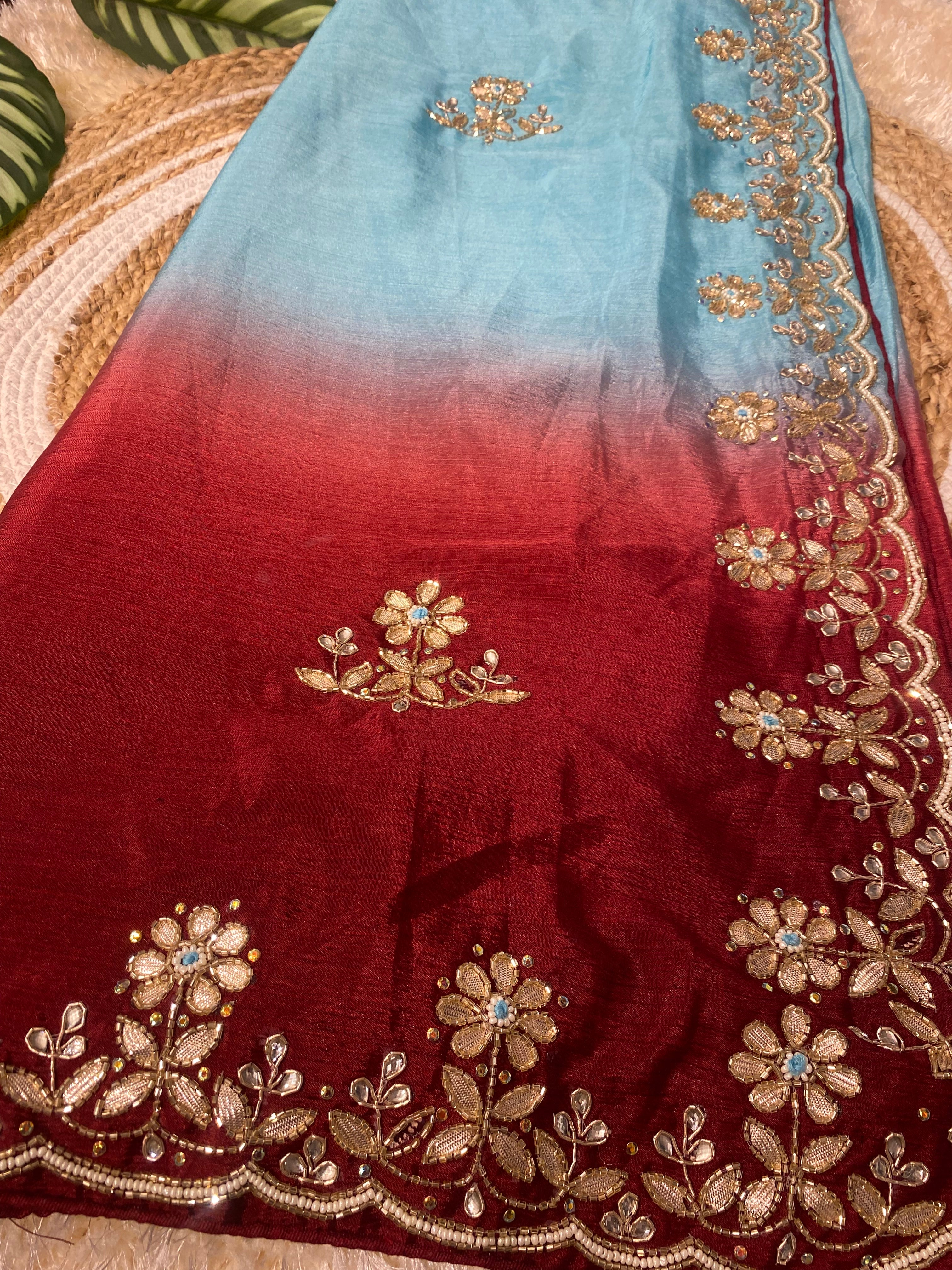 Close-up of an ombre saree blending from sky blue to deep red, featuring intricate gold floral embroidery with small blue stone accents and a scalloped embroidered border, elegantly displayed on a woven jute mat with decorative green leaves. ✨