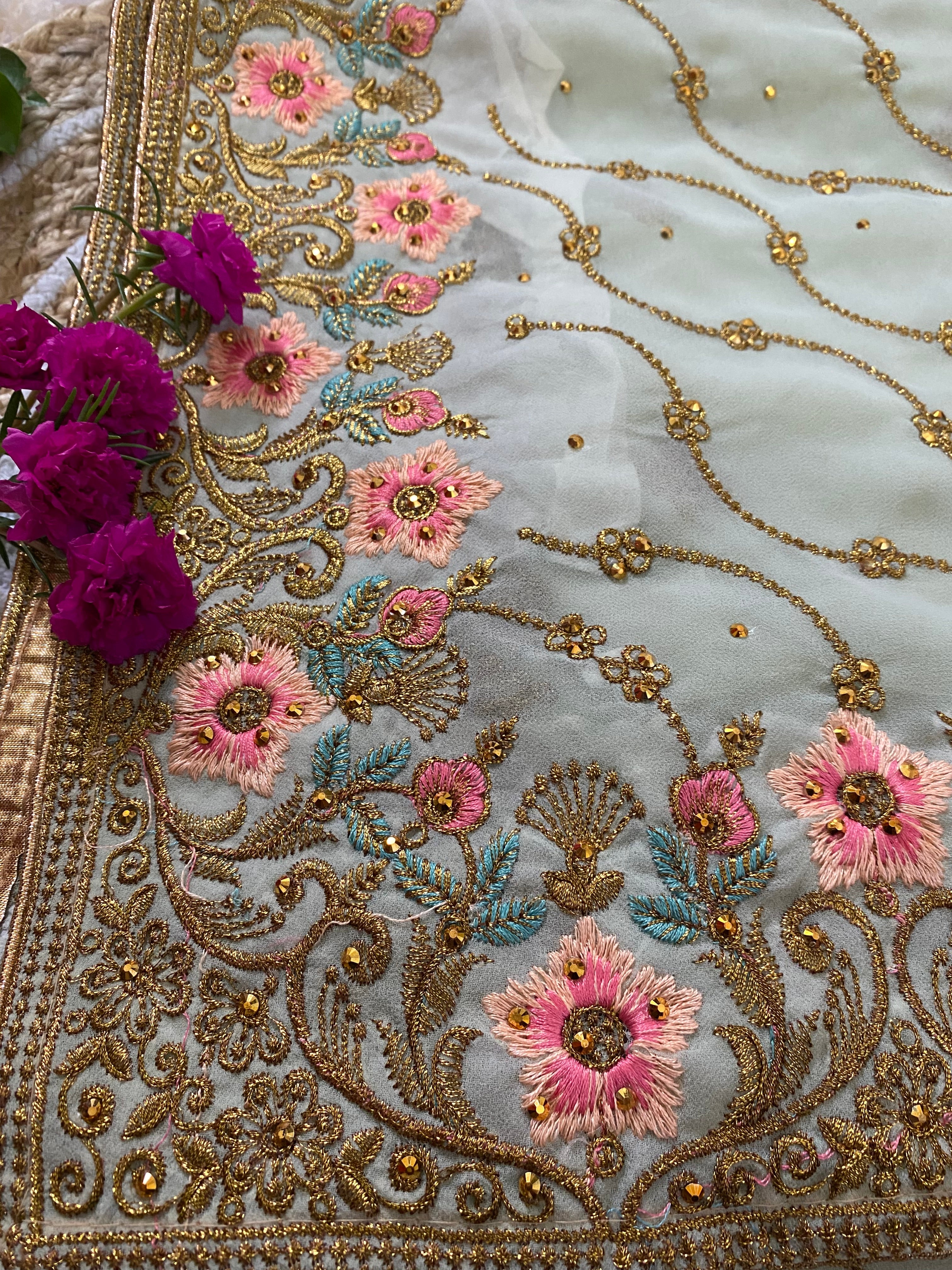 Close-up of a sheer pastel mint saree featuring intricate gold thread embroidery with pink floral motifs, teal leaf accents, and delicate vine patterns, styled with bright pink flowers along the ornate embroidered border.