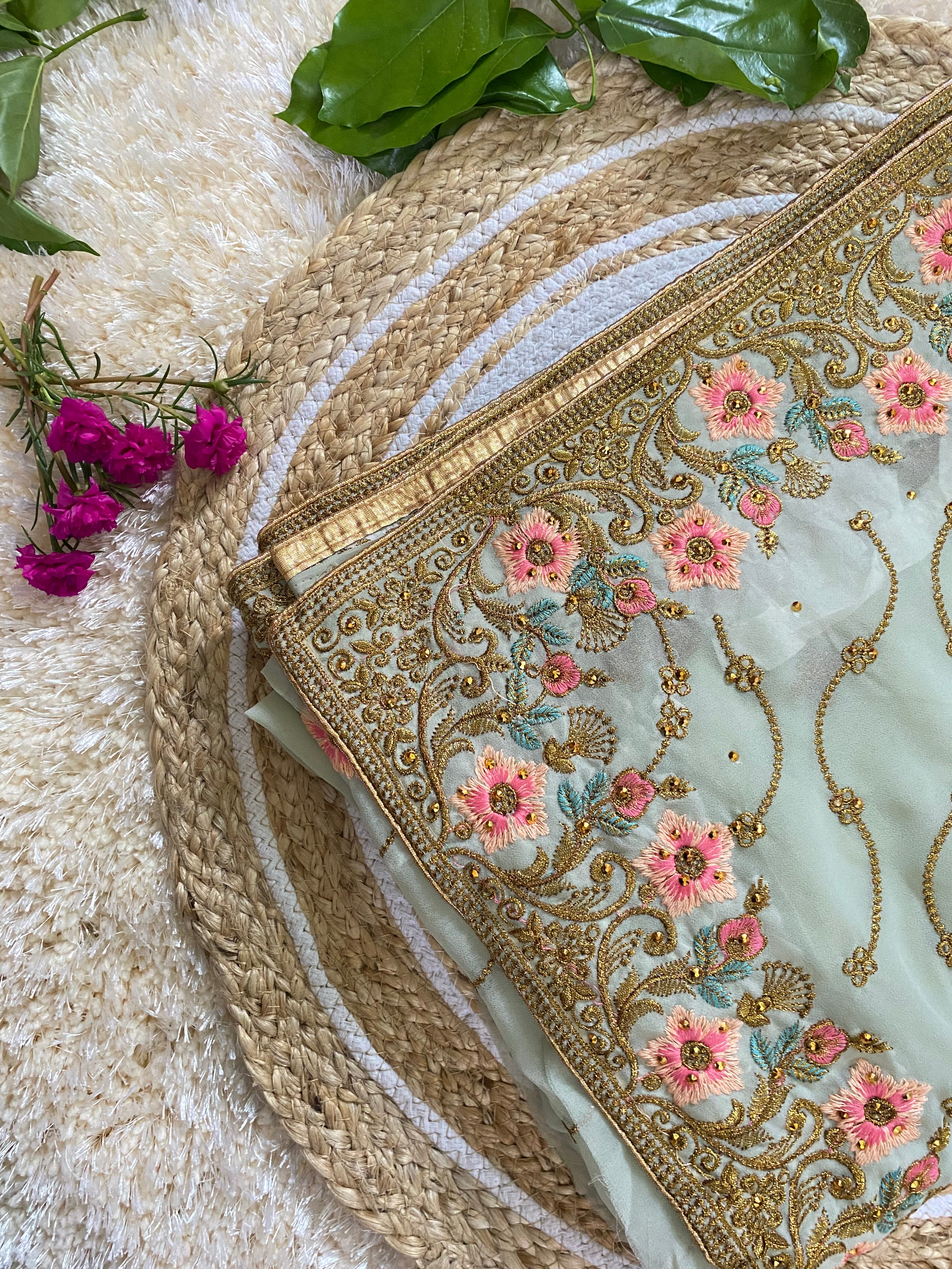 Folded pastel mint green saree with ornate gold thread embroidery, featuring pink floral motifs, teal leaf accents, and delicate vine patterns along the border, styled on a woven jute mat with green leaves and bright pink flowers. ✨