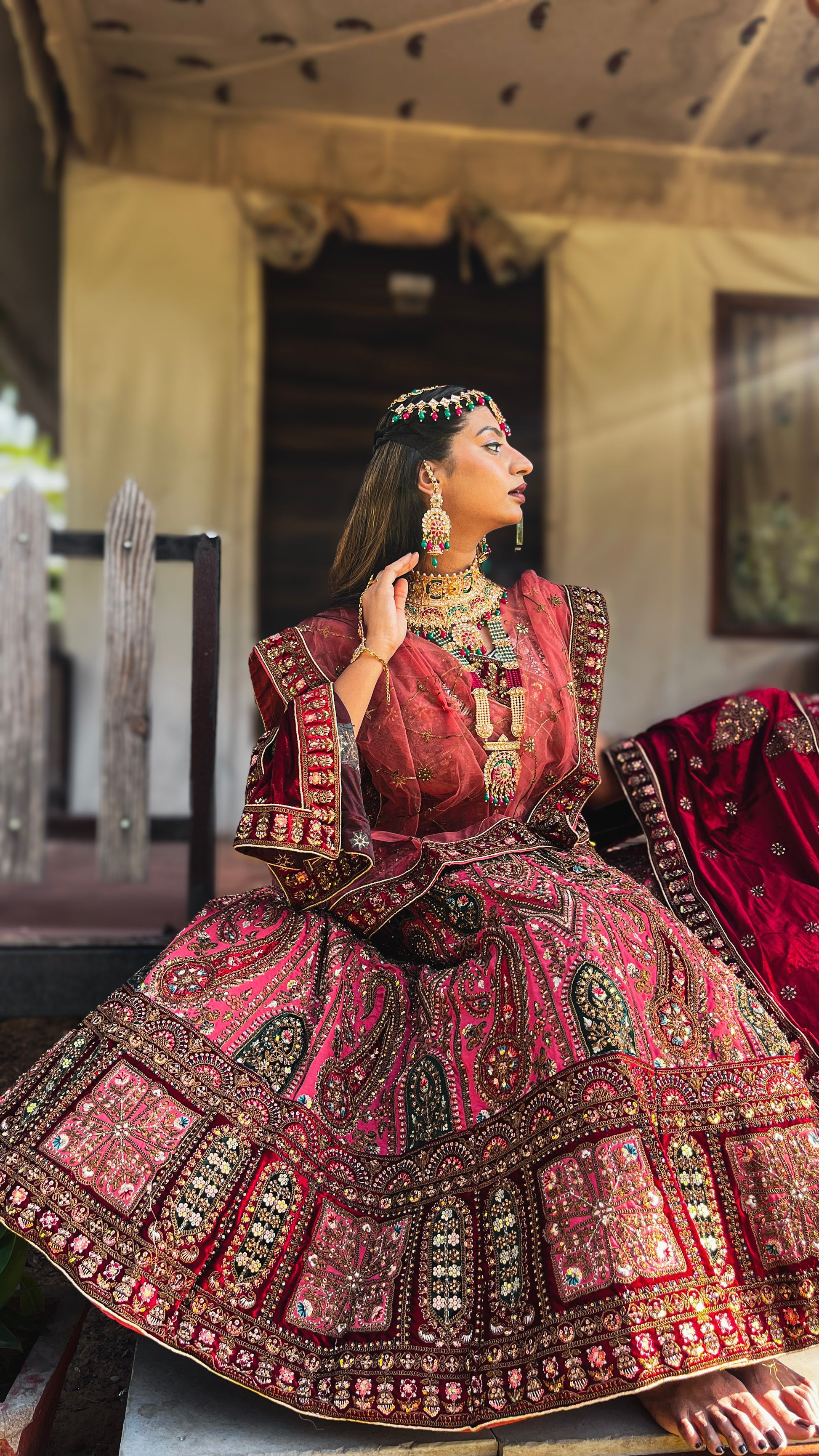 A model wearing Amai Jaipur’s intricately handcrafted red Antriya bridal lehenga, embellished with heavy zari, gota patti, and traditional Rajasthani embroidery, styled with ornate gold and multicolor gemstone jewelry, captured in a royal outdoor setting.