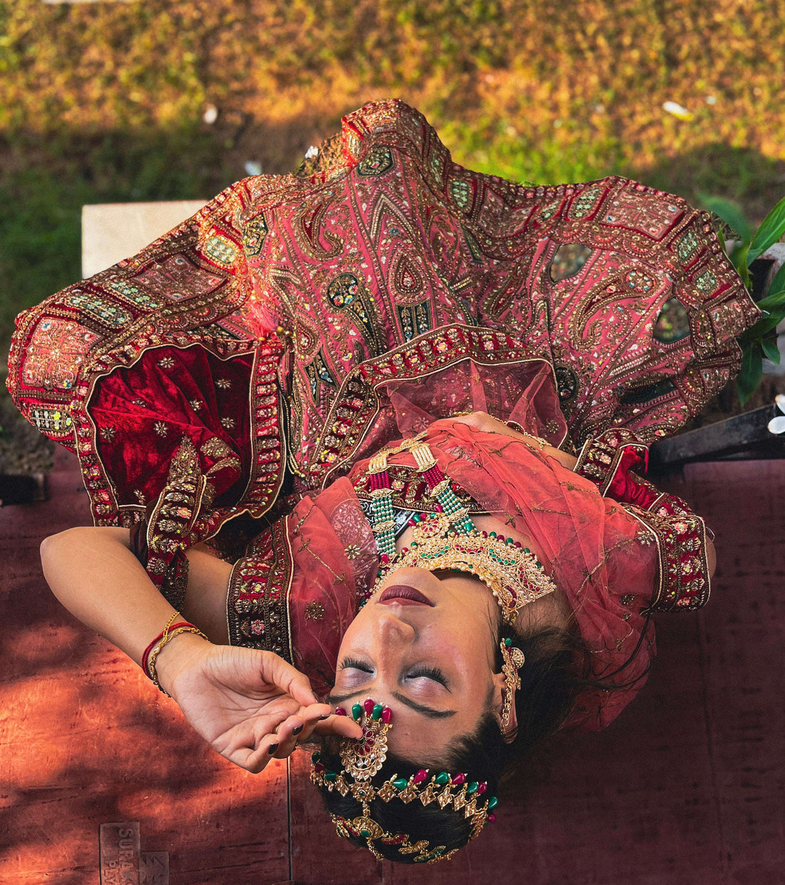 Model wearing Amai Jaipur’s Antriya Lehenga in red and gold, featuring heavy hand-embroidered Zari, mirror, and Kardhana work, captured in a creative overhead pose highlighting intricate bridal craftsmanship and traditional Indian design.