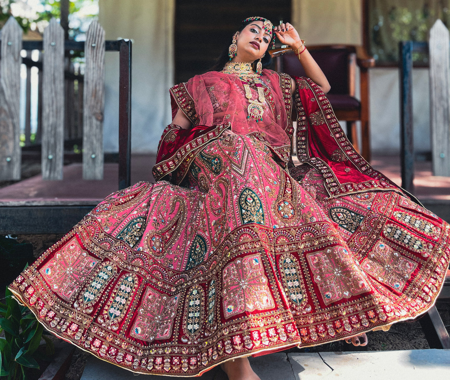 Model wearing Amai Jaipur’s ‘Antriya’ bridal lehenga in rich red with heavy hand-embroidery, mirror work, and traditional Indian craftsmanship, showcased in a seated pose highlighting the intricate flare and regal detailing.